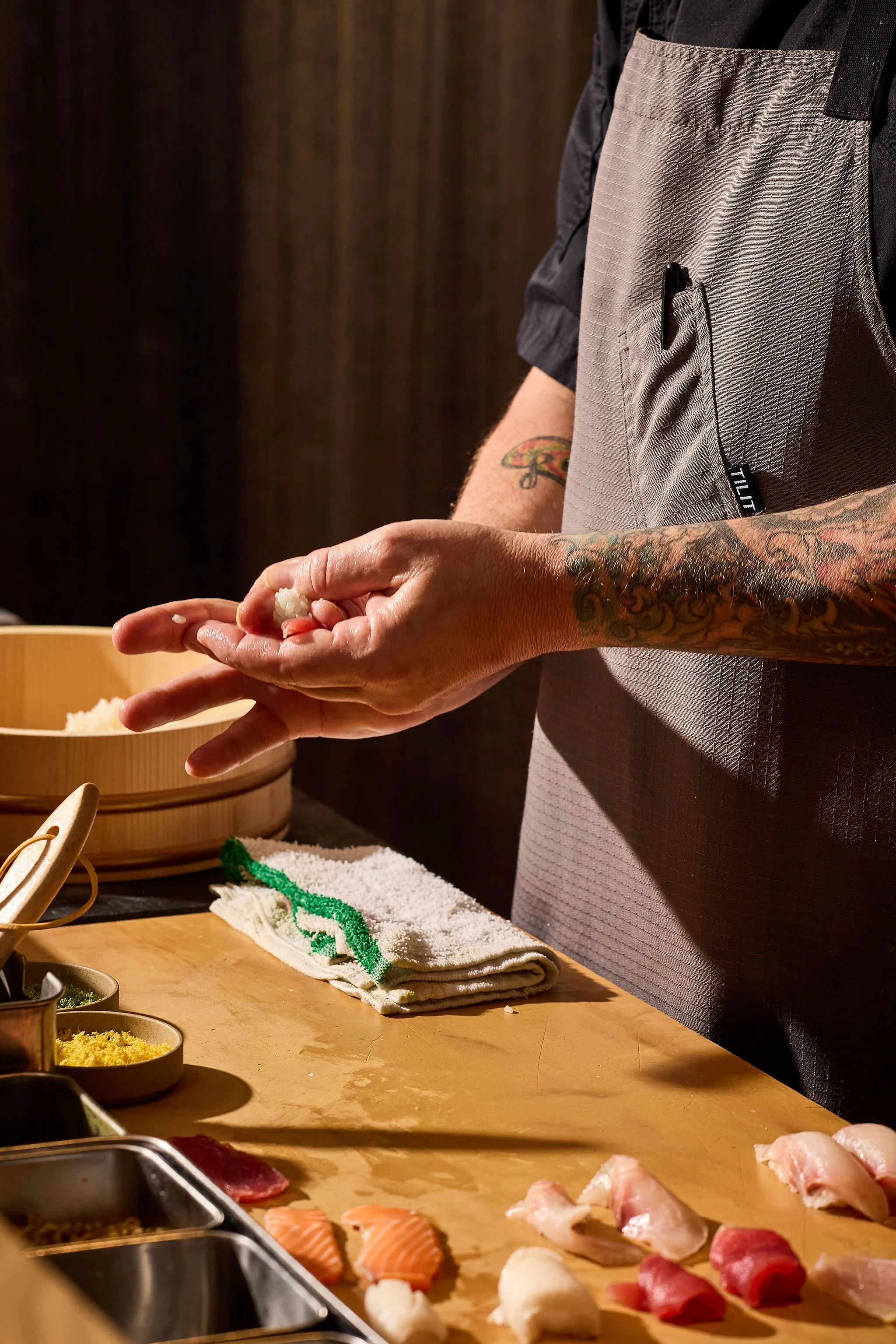 A chef with tattoos rolling sushi at a wooden counter, with various sushi ingredients and tools visible.