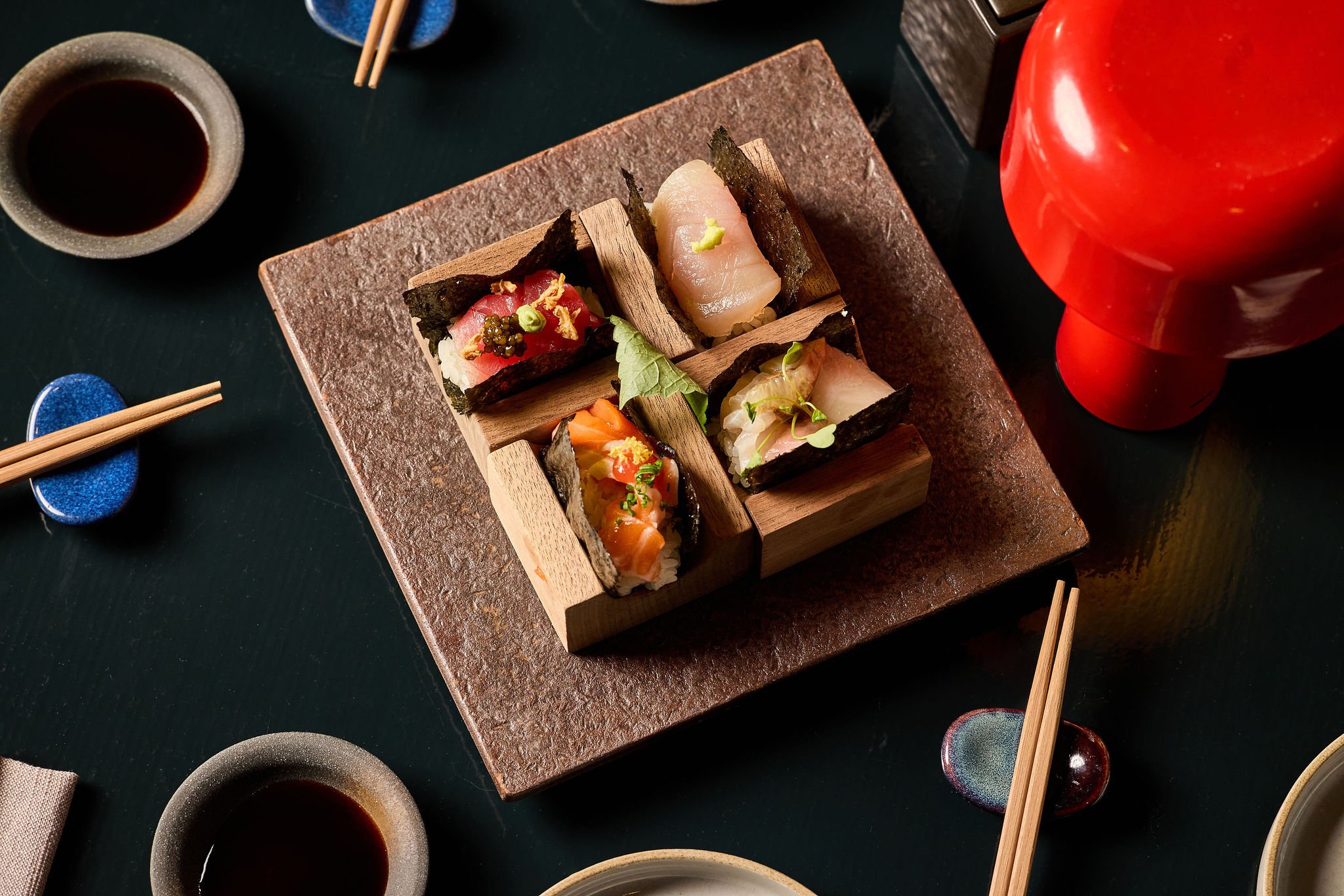 Assorted sushi pieces on a wooden serving block with soy sauce, chopsticks, and a red teapot on a black table.