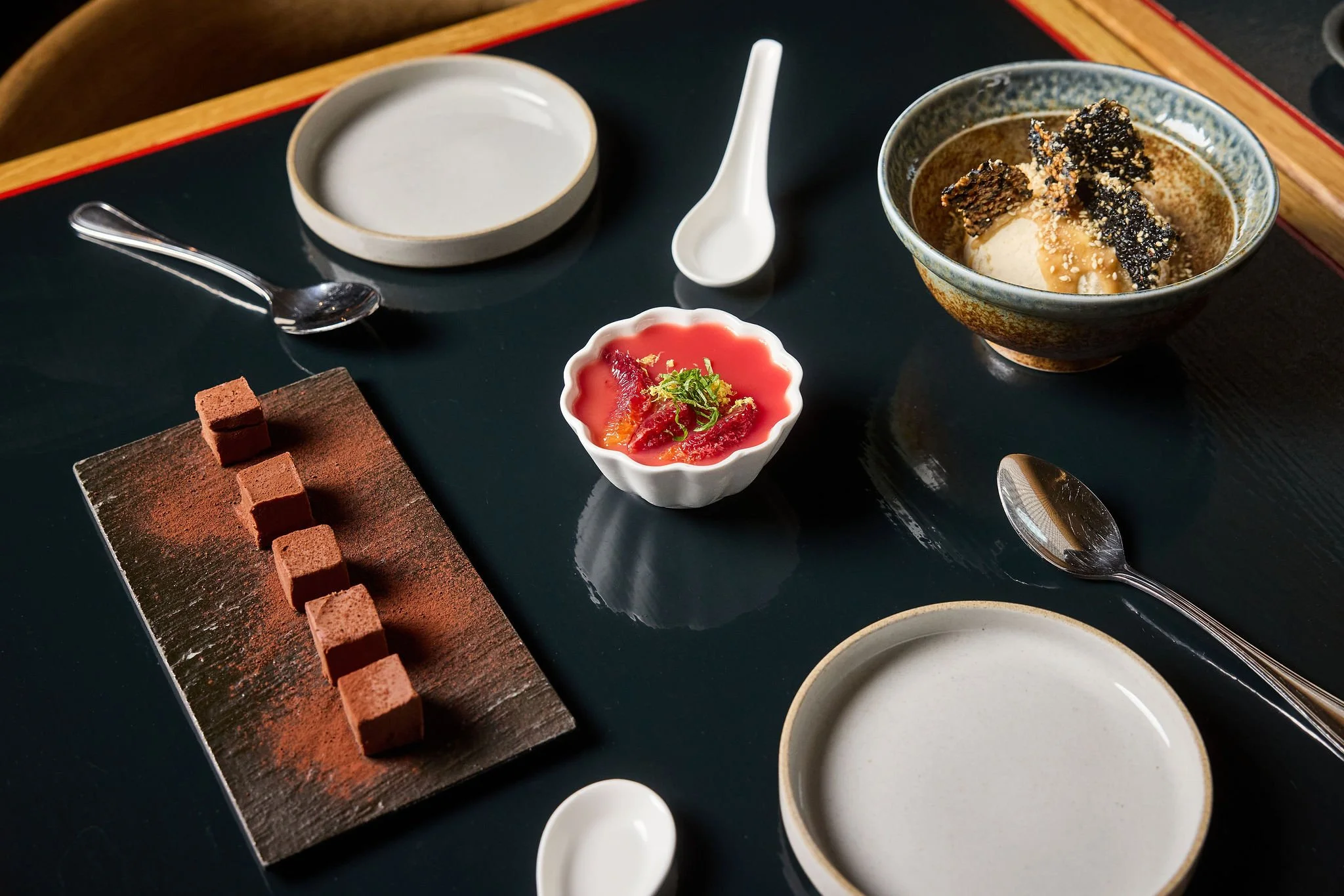 Assorted Japanese desserts on a black table, including a row of chocolate truffles, a small dish of red fruit jelly, a bowl of ice cream with toppings, and empty bowls and spoons.
