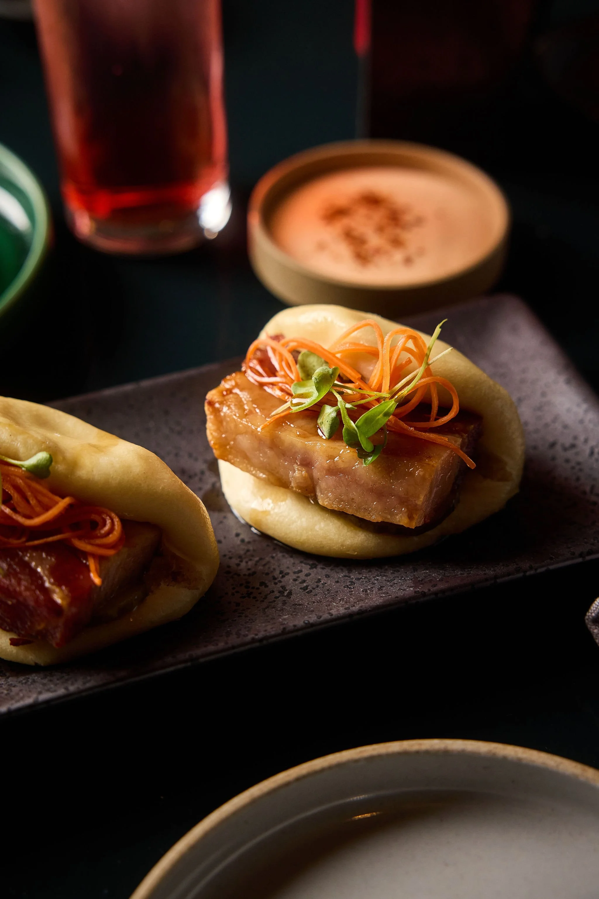Close-up of steamed pork buns topped with pork belly, julienned carrots, and microgreens on a black rectangular plate, with a glass of dark soda and a small bowl of dipping sauce in the background.