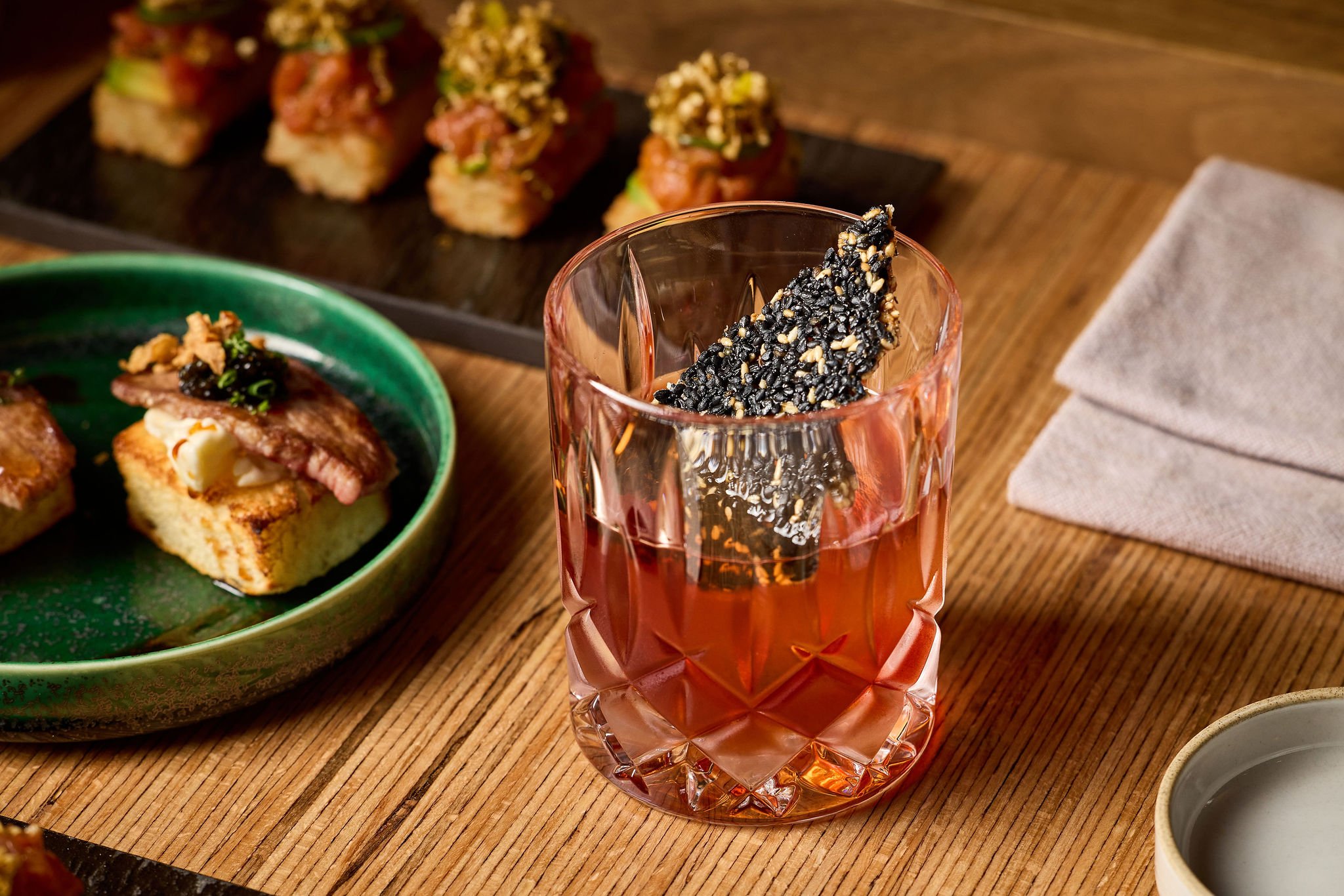 Glass of pink-colored beverage with a black sesame seaweed snack in it, surrounded by assorted sushi and Japanese dishes on a wooden table.