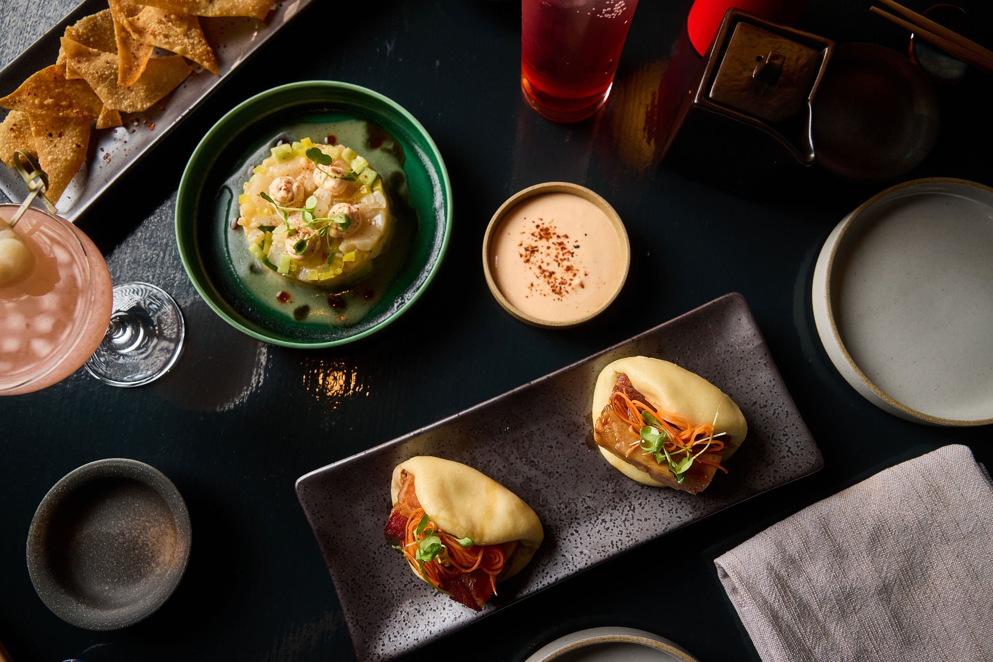 Assorted Asian dishes including spring rolls, dumpling soup, a pink cocktail, and dipping sauces on a black table.