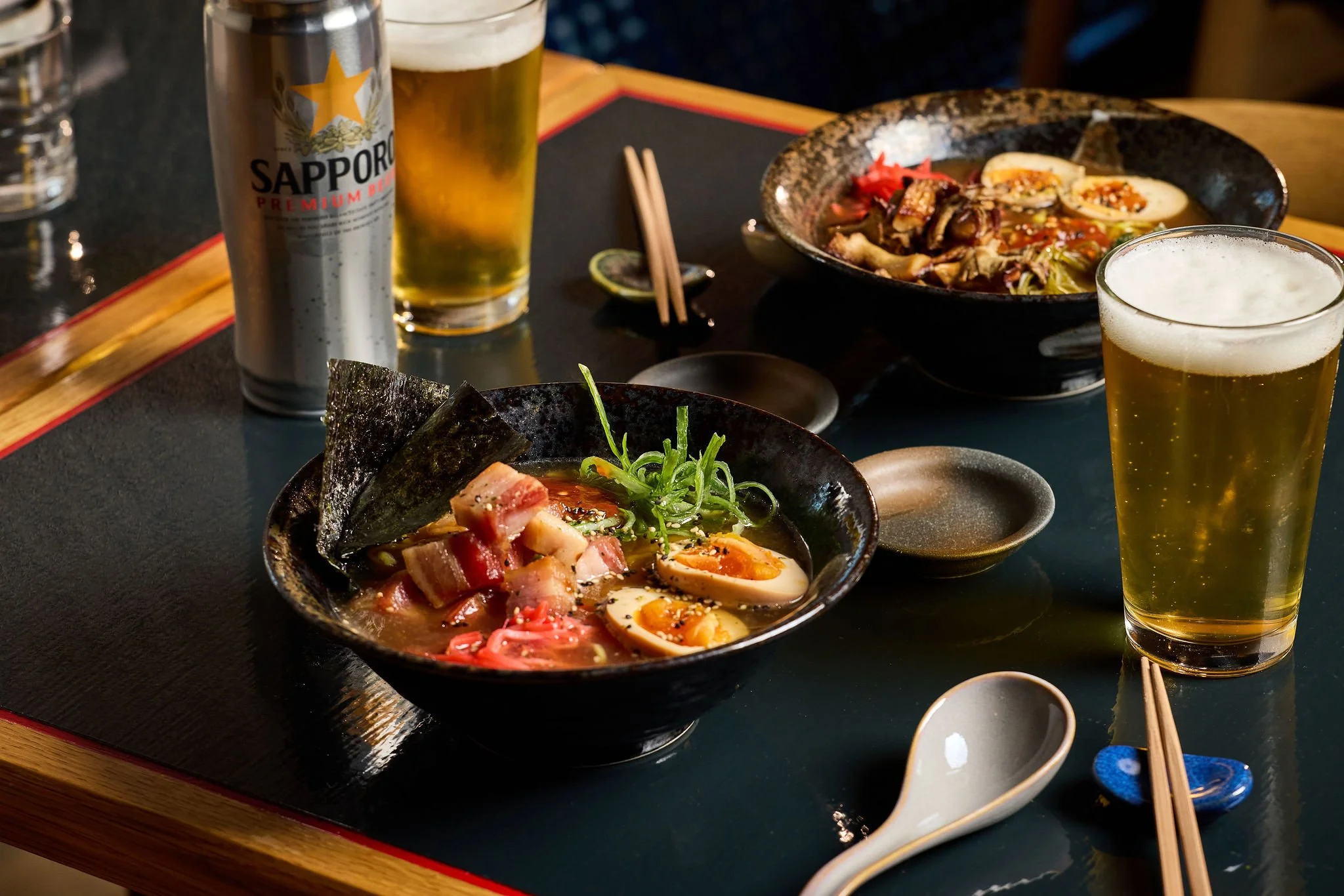 Two bowls of ramen with soft-boiled eggs, pork, seaweed, green onions, and vegetables, served on a dark table with glasses of beer, chopsticks, and spoons.