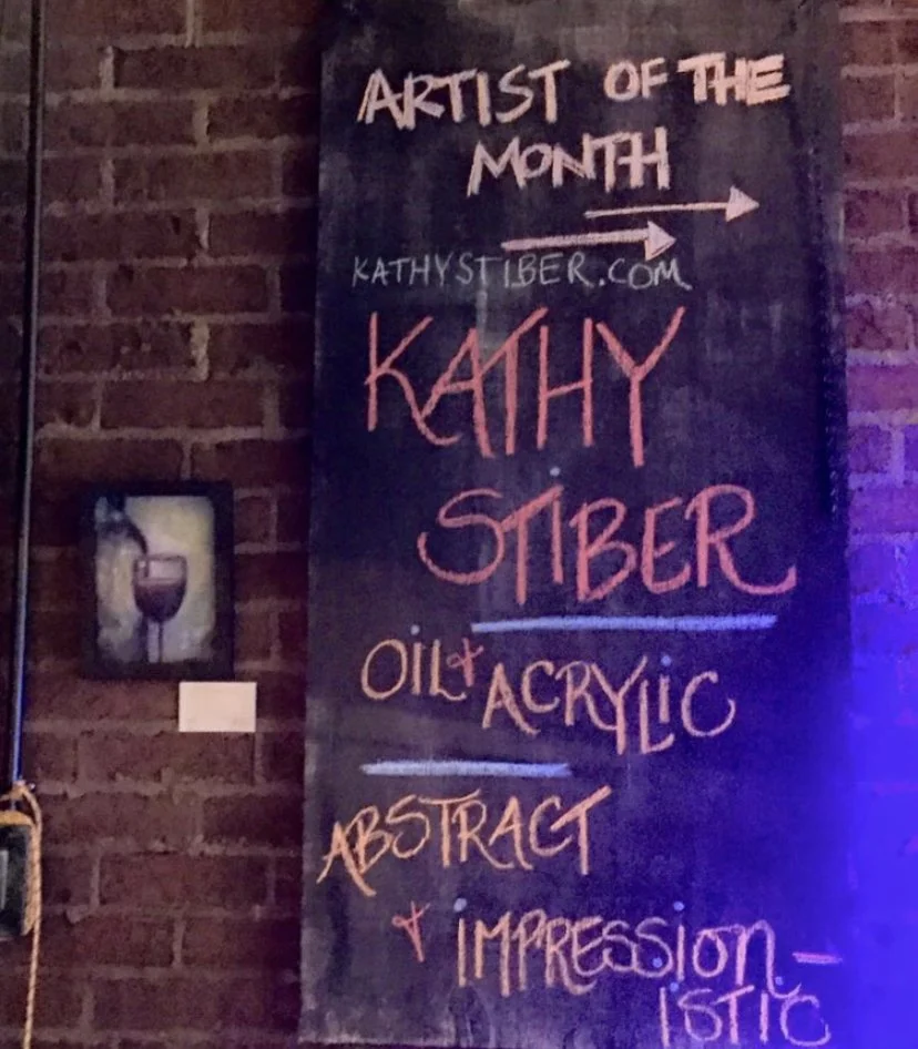 A black chalkboard sign with colorful handwritten text announcing Kathy Siber as the artist of the month, advertising her work in oil and acrylic, emphasizing abstract and impressionistic styles. The sign is hung on a brick wall.