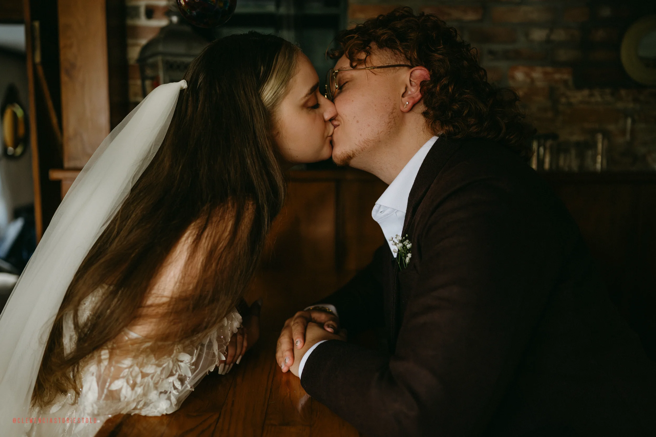 A bride and groom share a kiss at a rustic wedding venue, leaning over a wooden table with hands clasped.