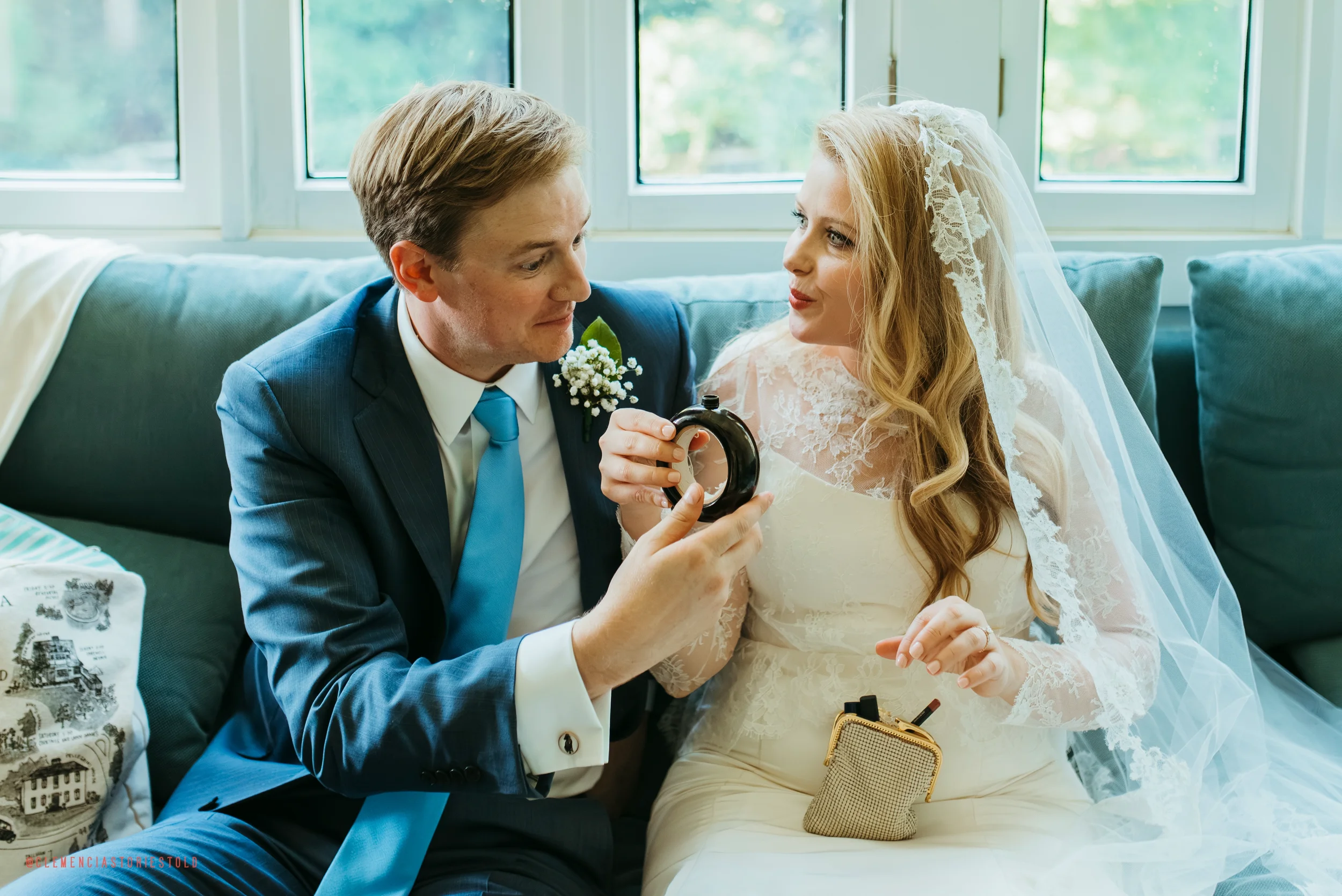 A groom in a blue suit and a bride in a white wedding dress sitting on a sofa, holding a black ring bottle, with the bride wearing a veil and lace dress, in front of large windows.