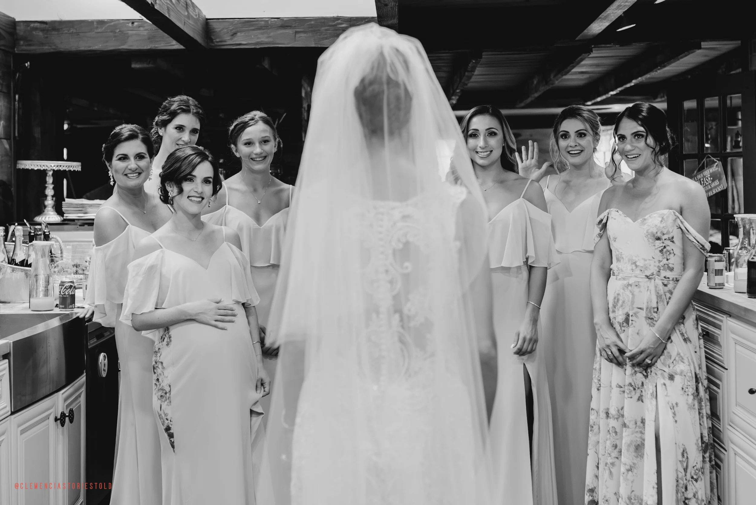 A bride in a wedding dress stands with her back to the camera, facing a group of bridesmaids and a woman, possibly her mother, in a kitchen or bar area. The bridesmaids wear matching light-colored dresses, and everyone is smiling or looking at the br