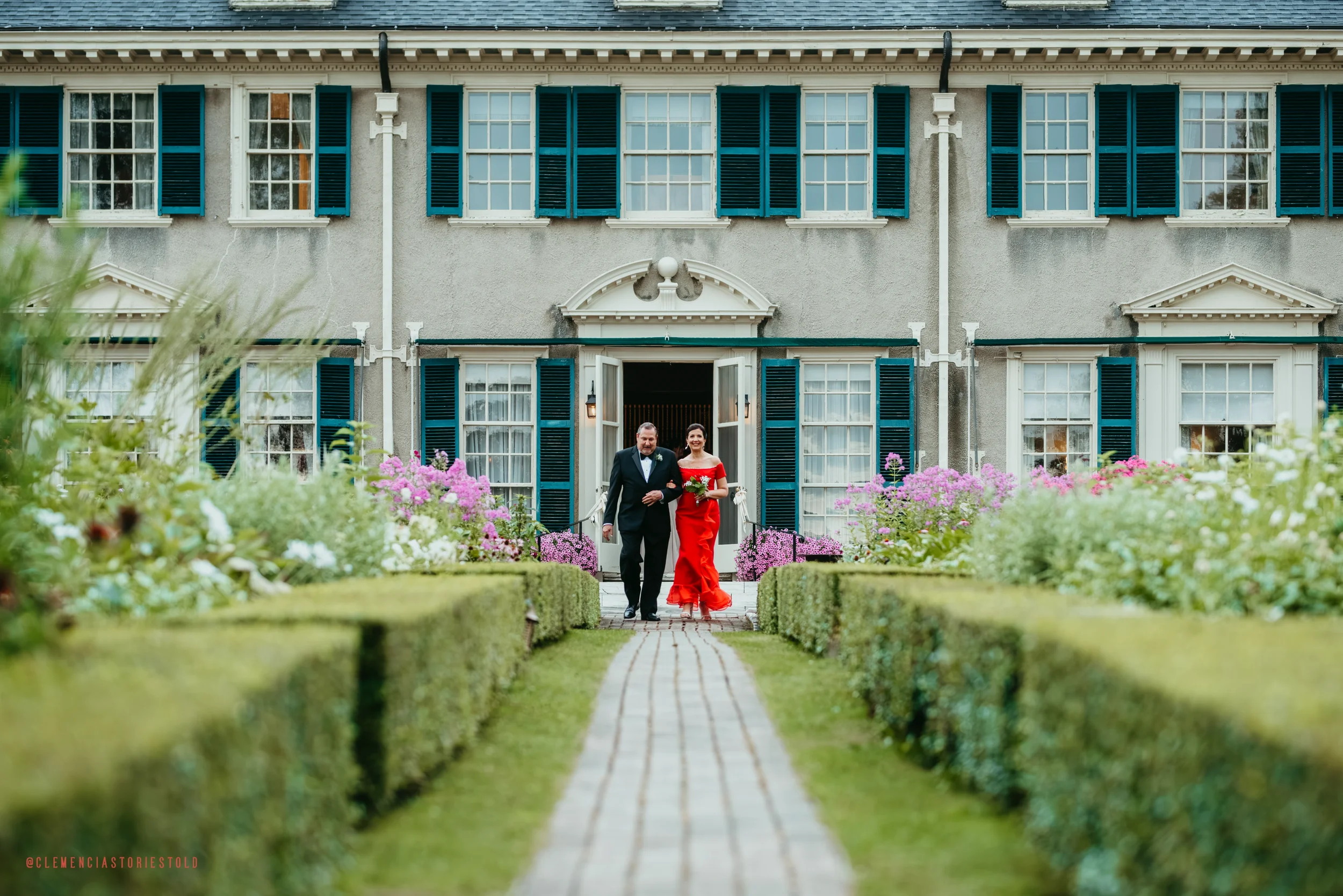 A bride in a red wedding dress and a groom in a black tuxedo walking down a garden pathway towards a large gray mansion with white trim and blue shutters, surrounded by colorful flowers.