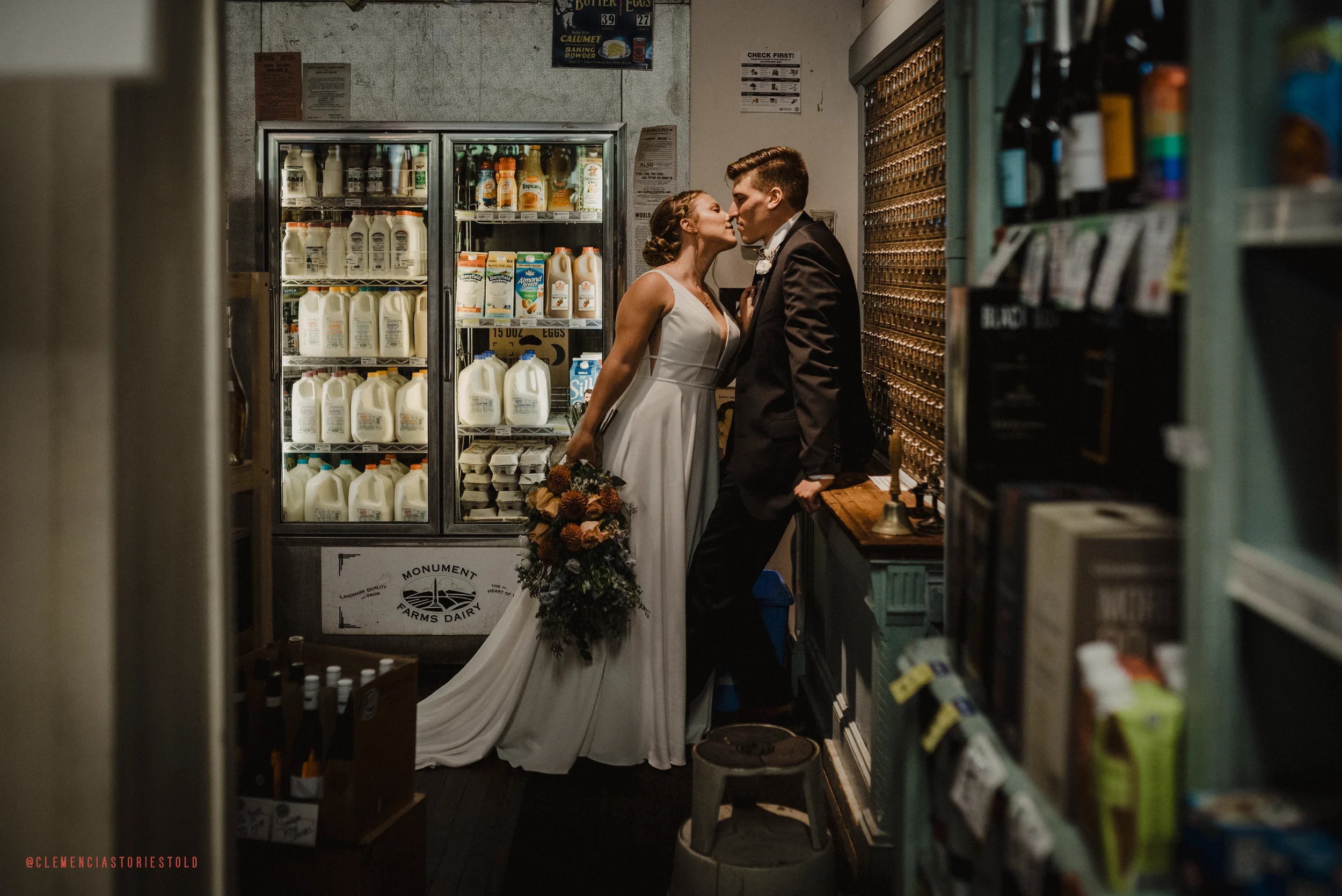 A bride and groom sharing an intimate moment in a small grocery store. The bride is holding a bouquet and wearing a white dress, while the groom is in a dark suit. They are standing close, about to kiss.