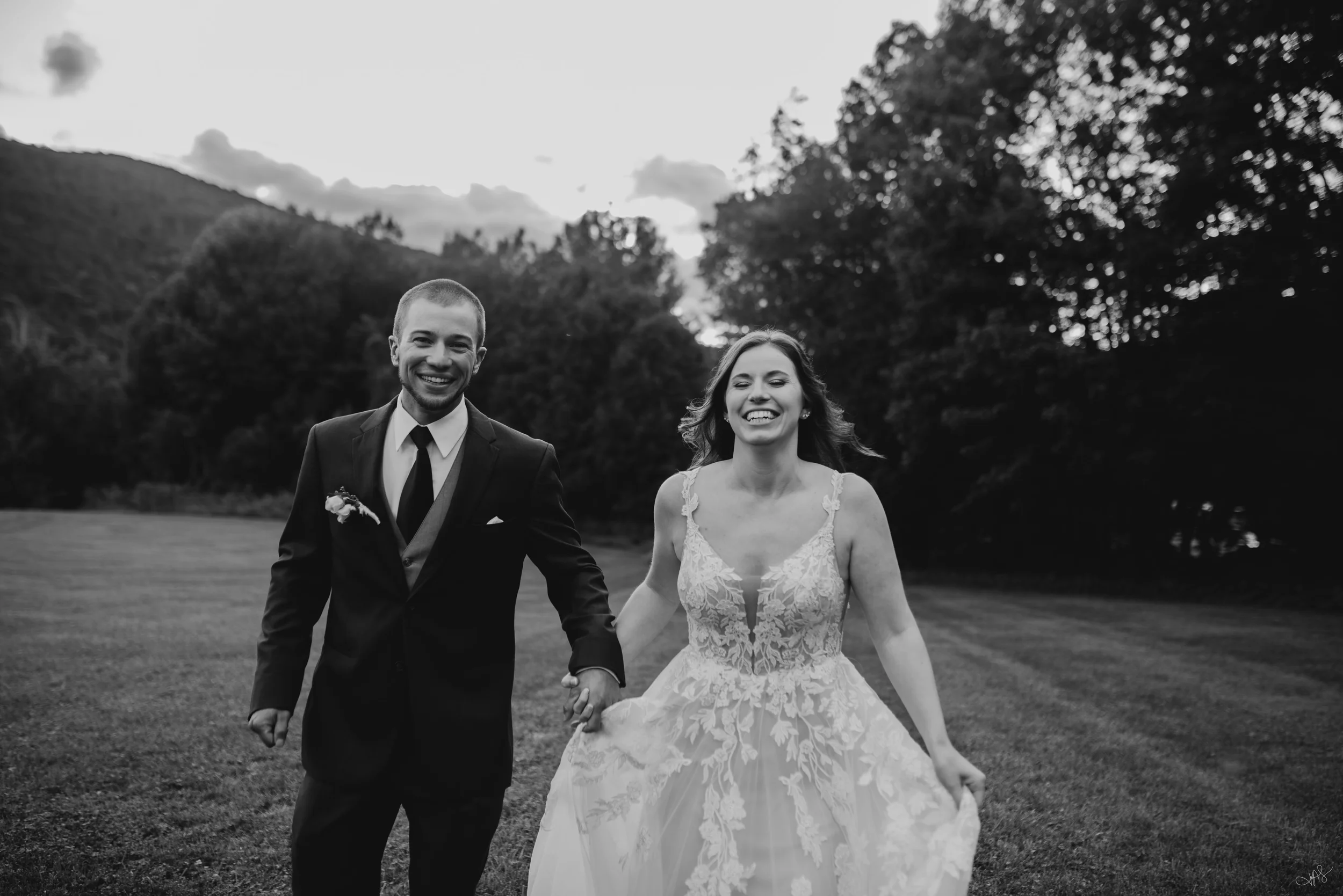 Black and white photo of a smiling young couple, a man in a suit and a woman in a lace wedding dress, walking hand in hand outdoors with trees and hills in the background.