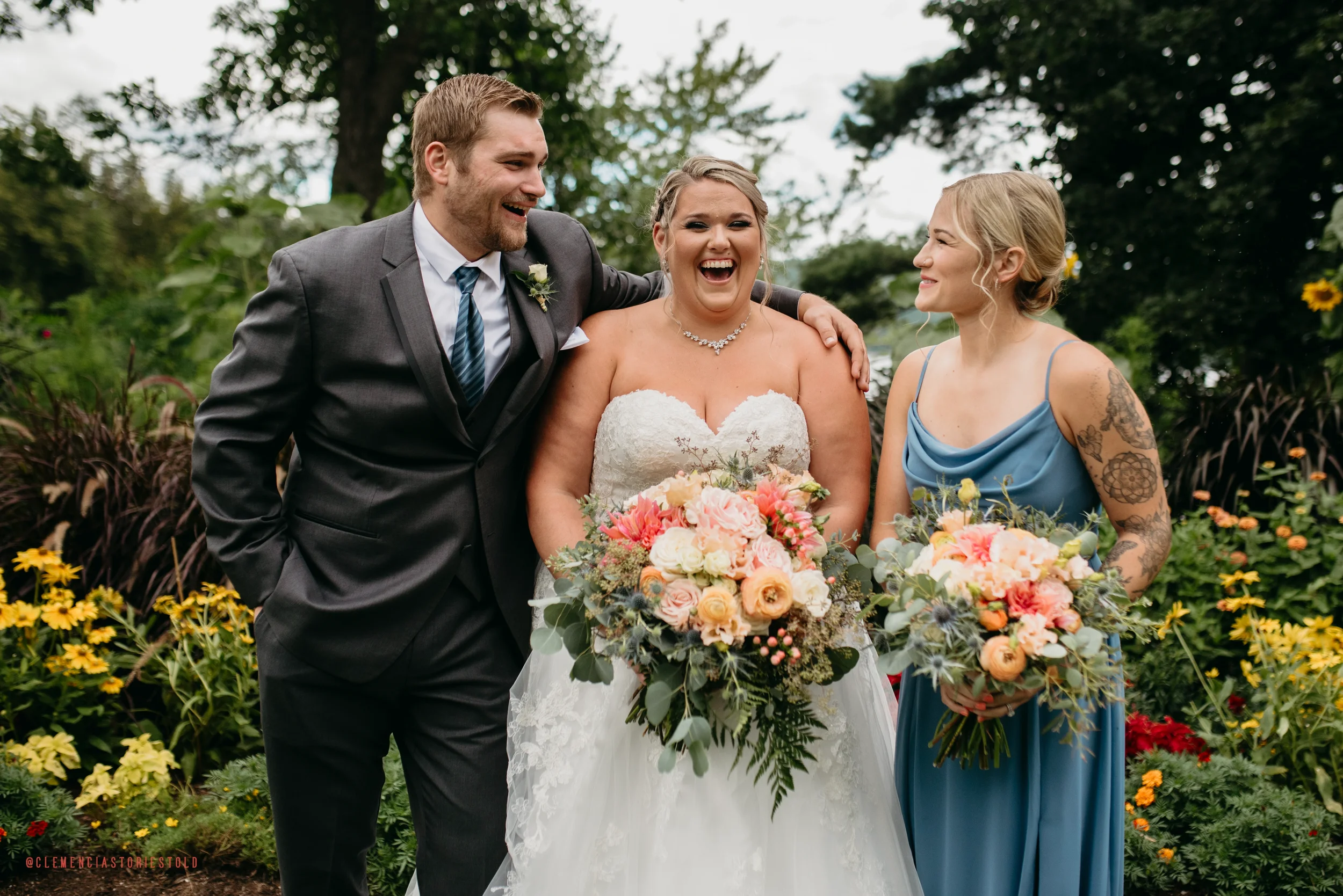 Three people, a bride in a white wedding dress holding a bouquet of flowers, a groom in a gray suit, and a bridesmaid in a blue dress, standing outdoors in a garden with green foliage and colorful flowers. The bride and groom are smiling and laughing