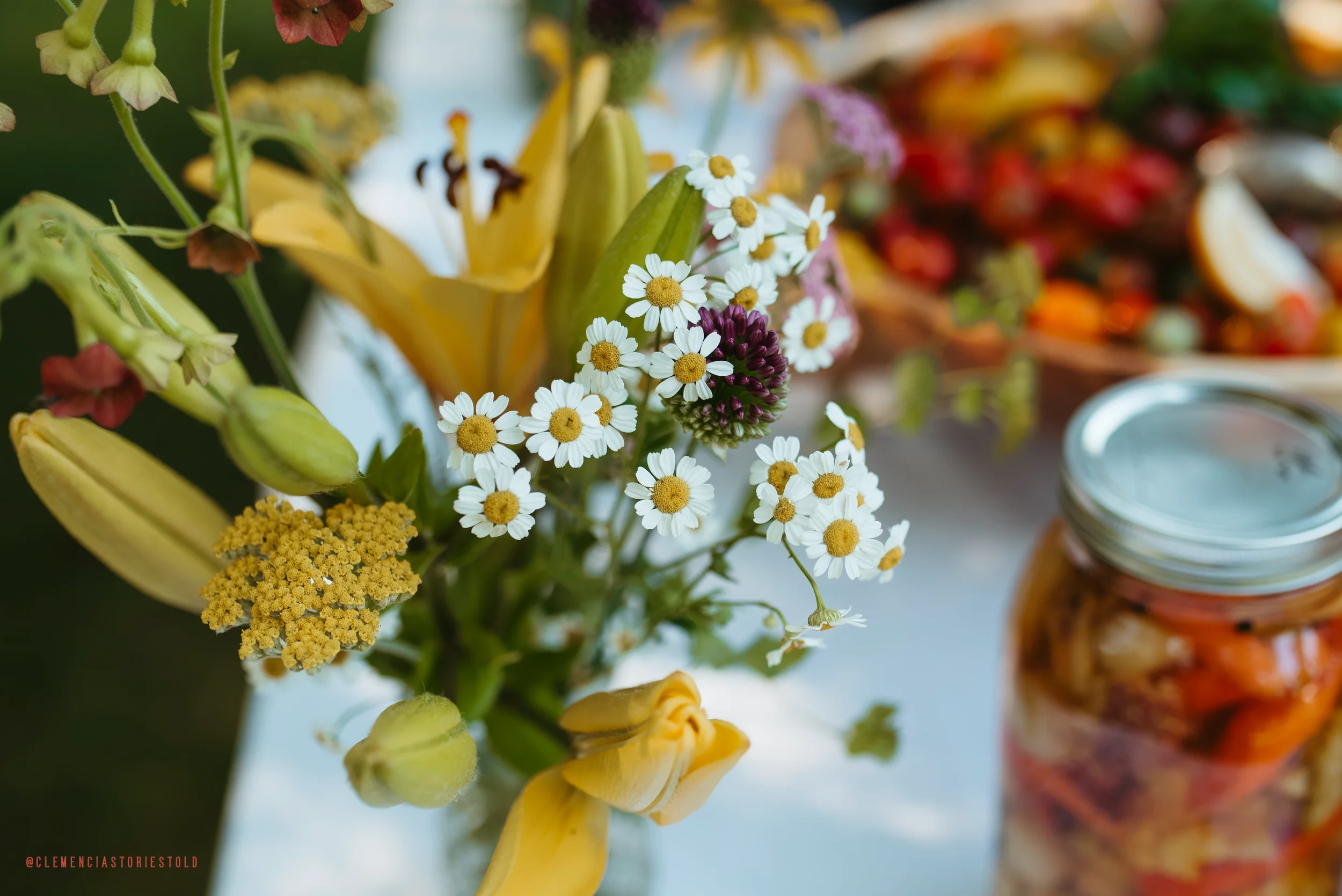 Close-up of a bouquet of assorted garden flowers, including daisies and lilies, with a jar of pickled vegetables nearby.
