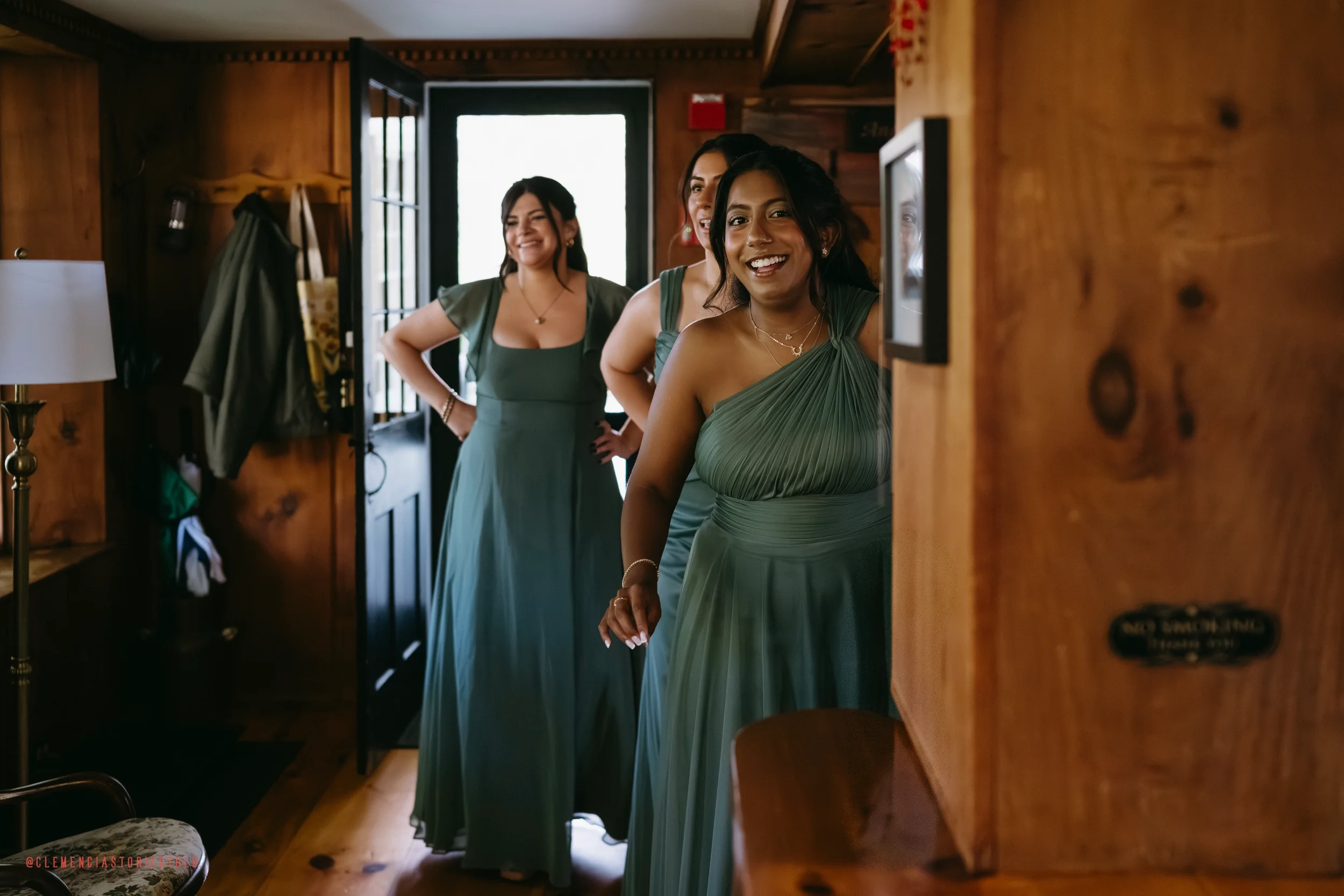 Four women in matching green dresses peering around a corner in a wooden room, smiling and enjoying the moment.