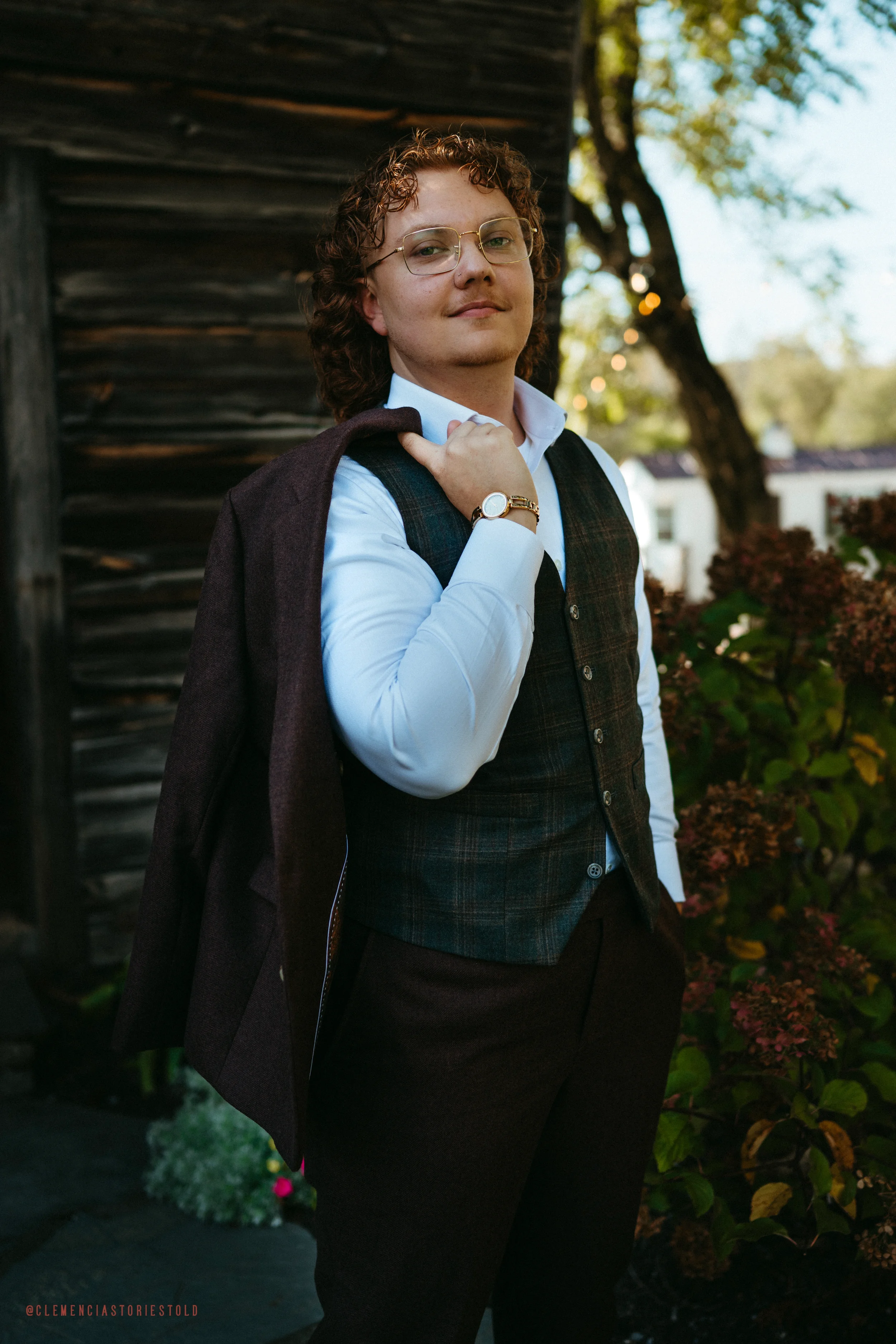 A young man with curly hair and glasses standing outdoors, wearing a white shirt, dark vest, and dark trousers, carrying a jacket over his shoulder, with a tree and plants in the background.