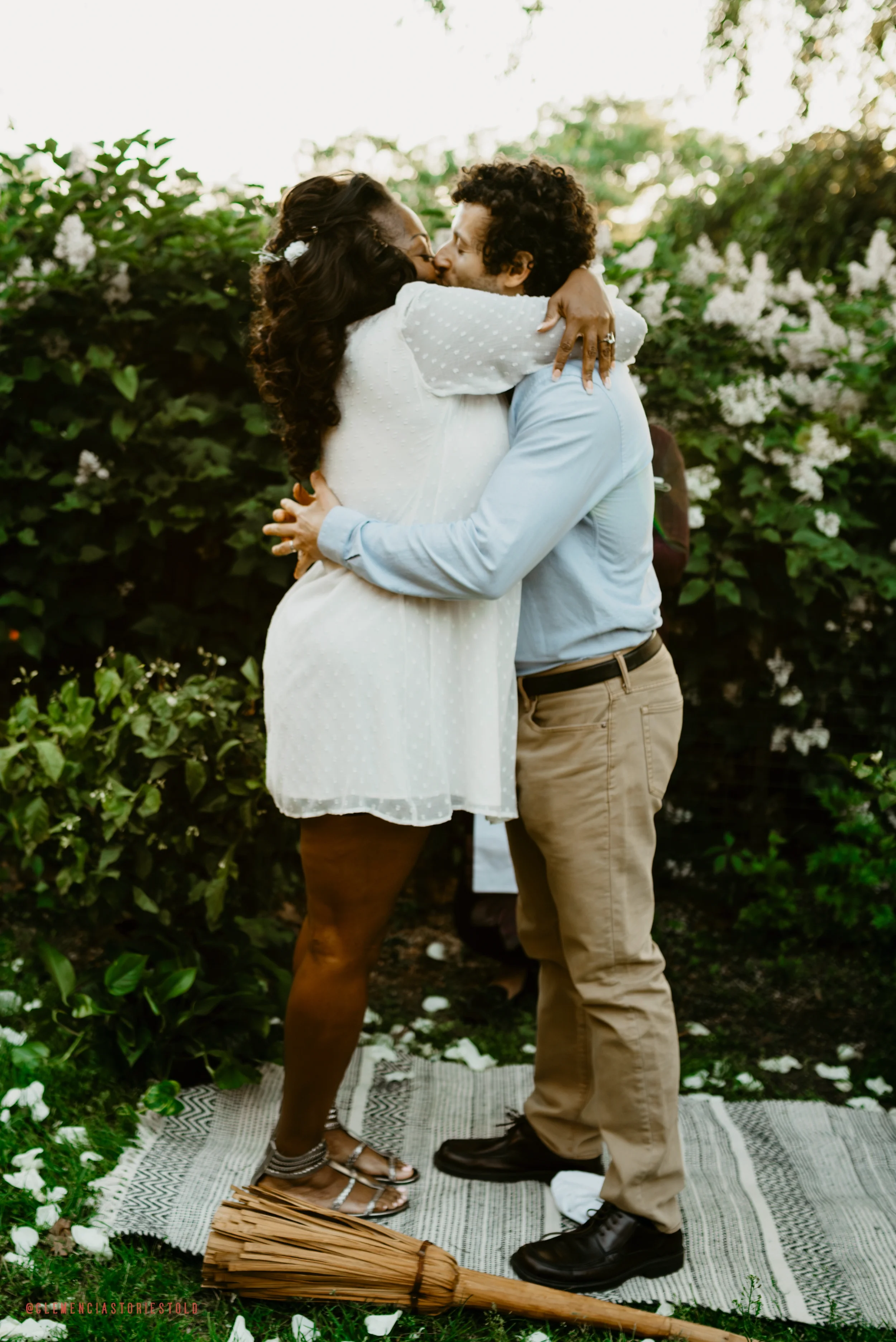 A couple sharing a kiss and hugging outdoors during a wedding ceremony, with trees and flowers in the background.