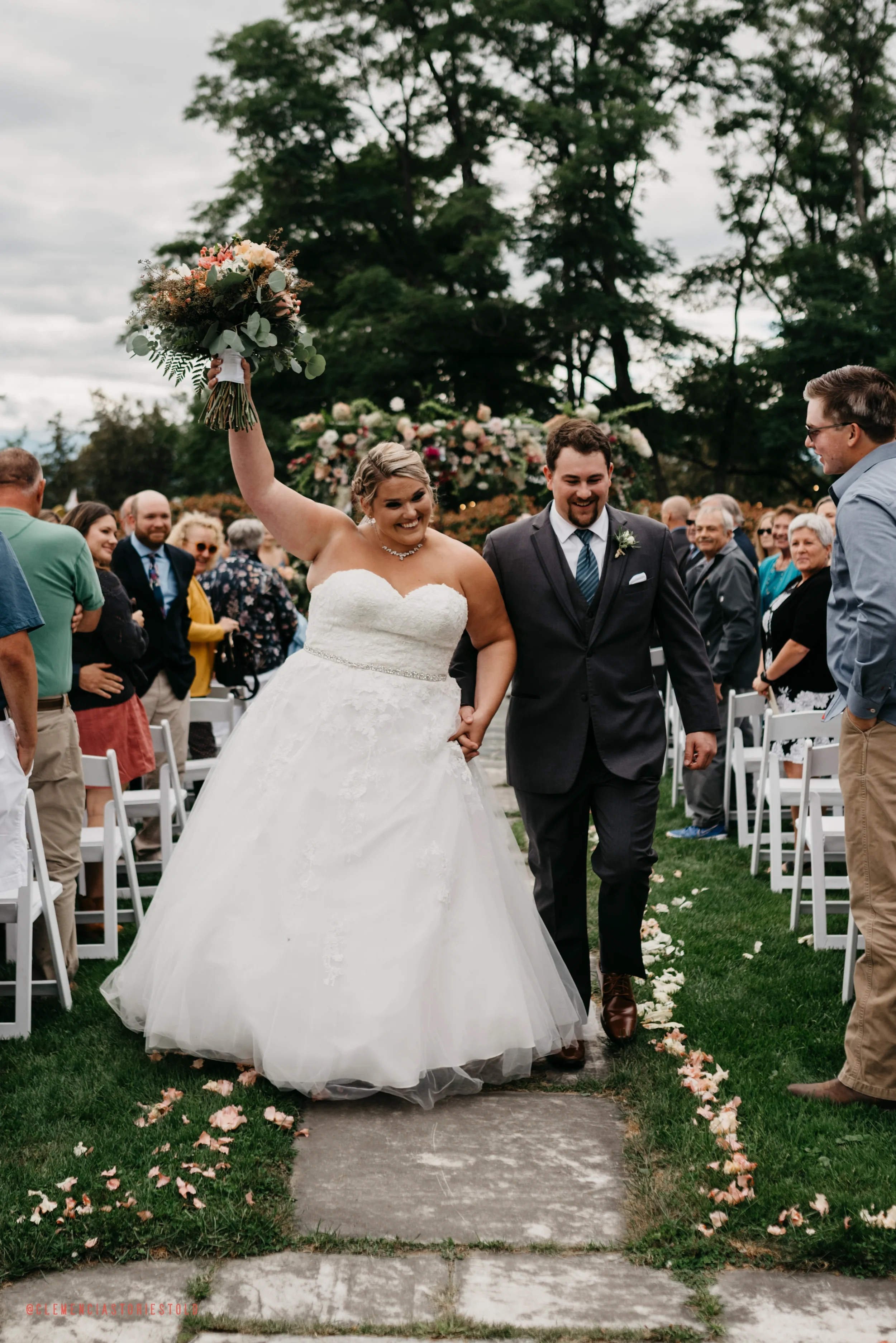 Bride in a white wedding gown holding a bouquet and smiling as she walks down the aisle with groom in a dark suit. Guests seated on both sides, trees and cloudy sky in the background.