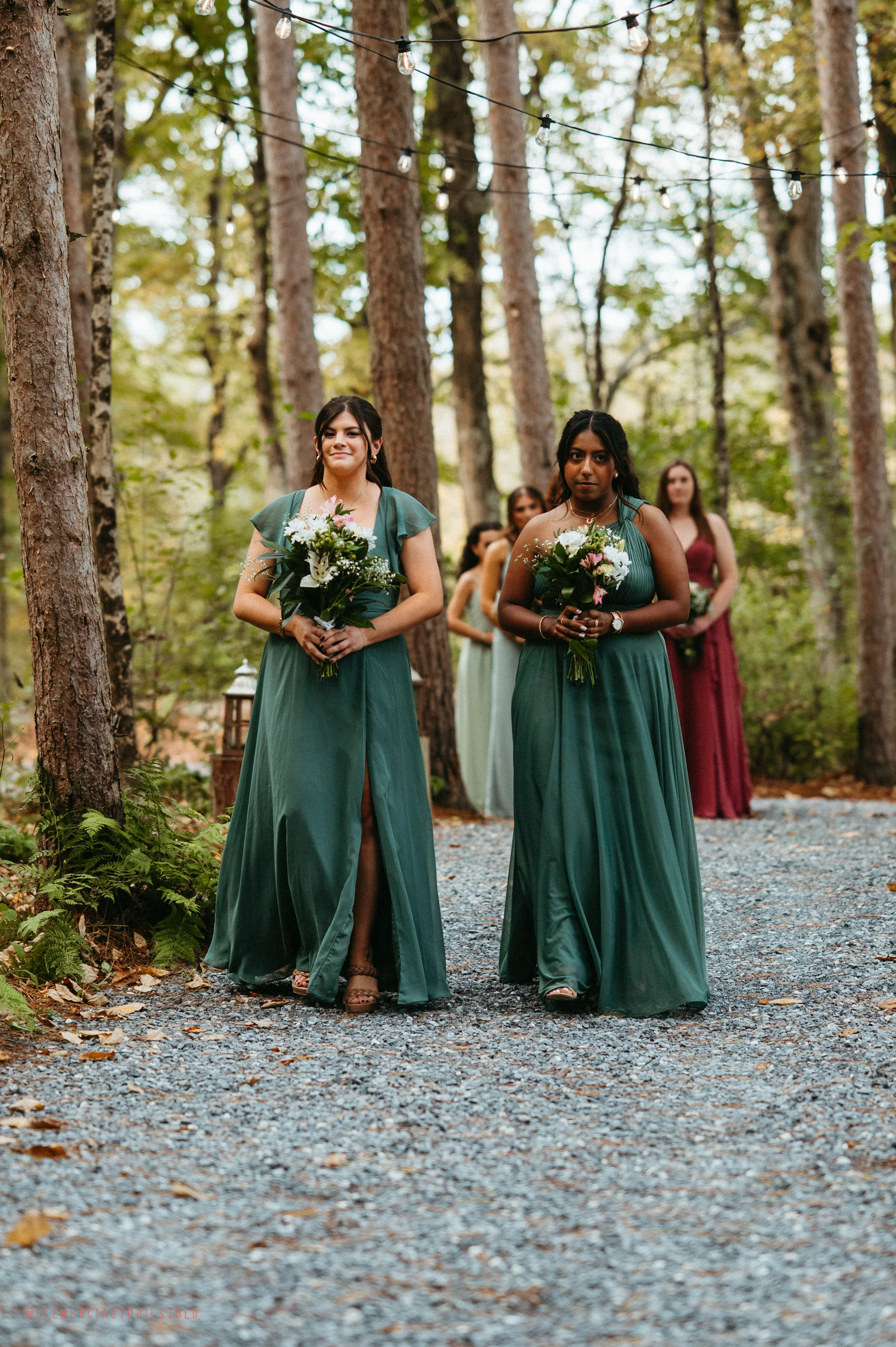 Group of women in long green dresses walking down a tree-lined outdoor aisle during a wedding ceremony, holding bouquets of flowers.
