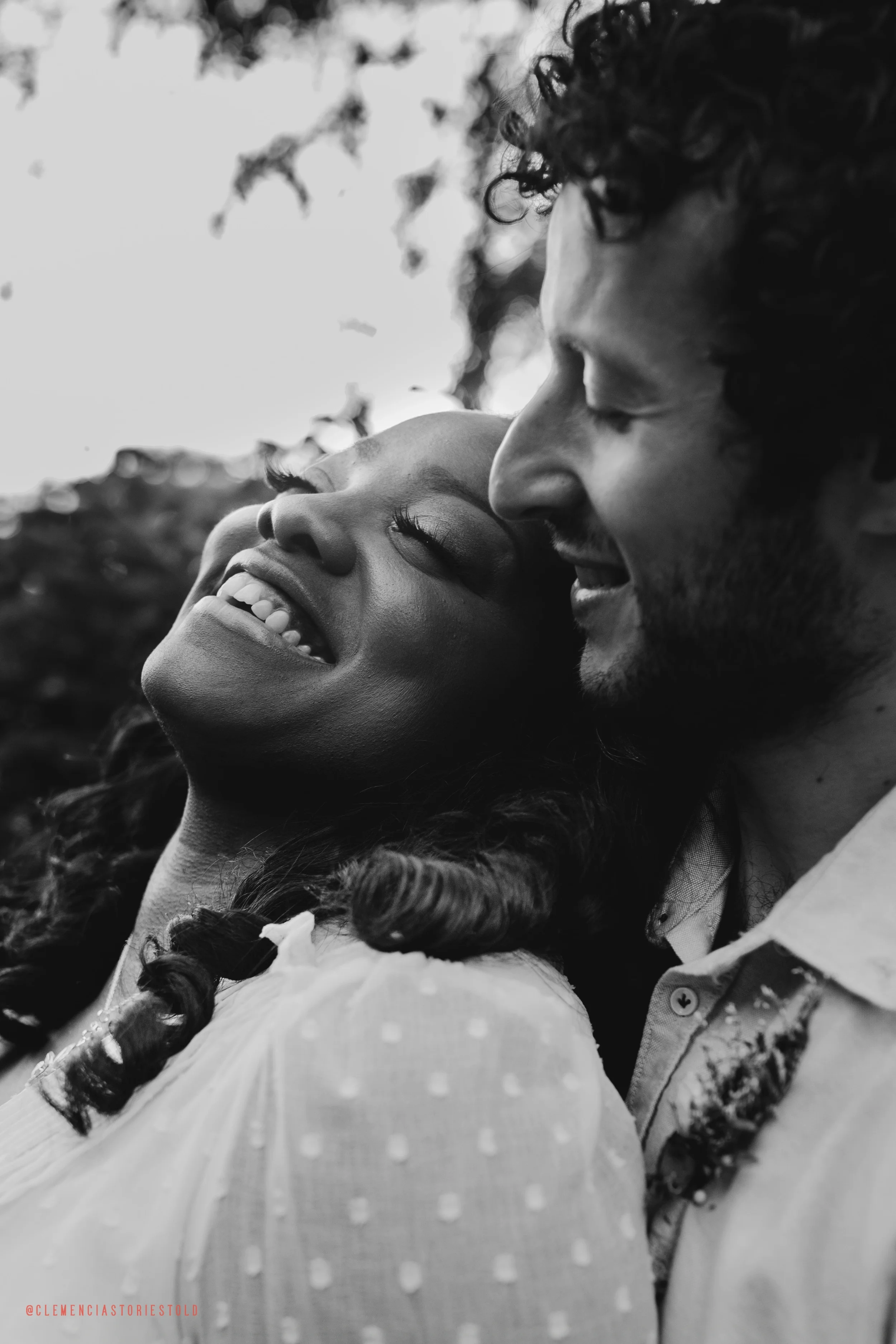 A smiling couple with closed eyes leaning close together outdoors, in black and white.