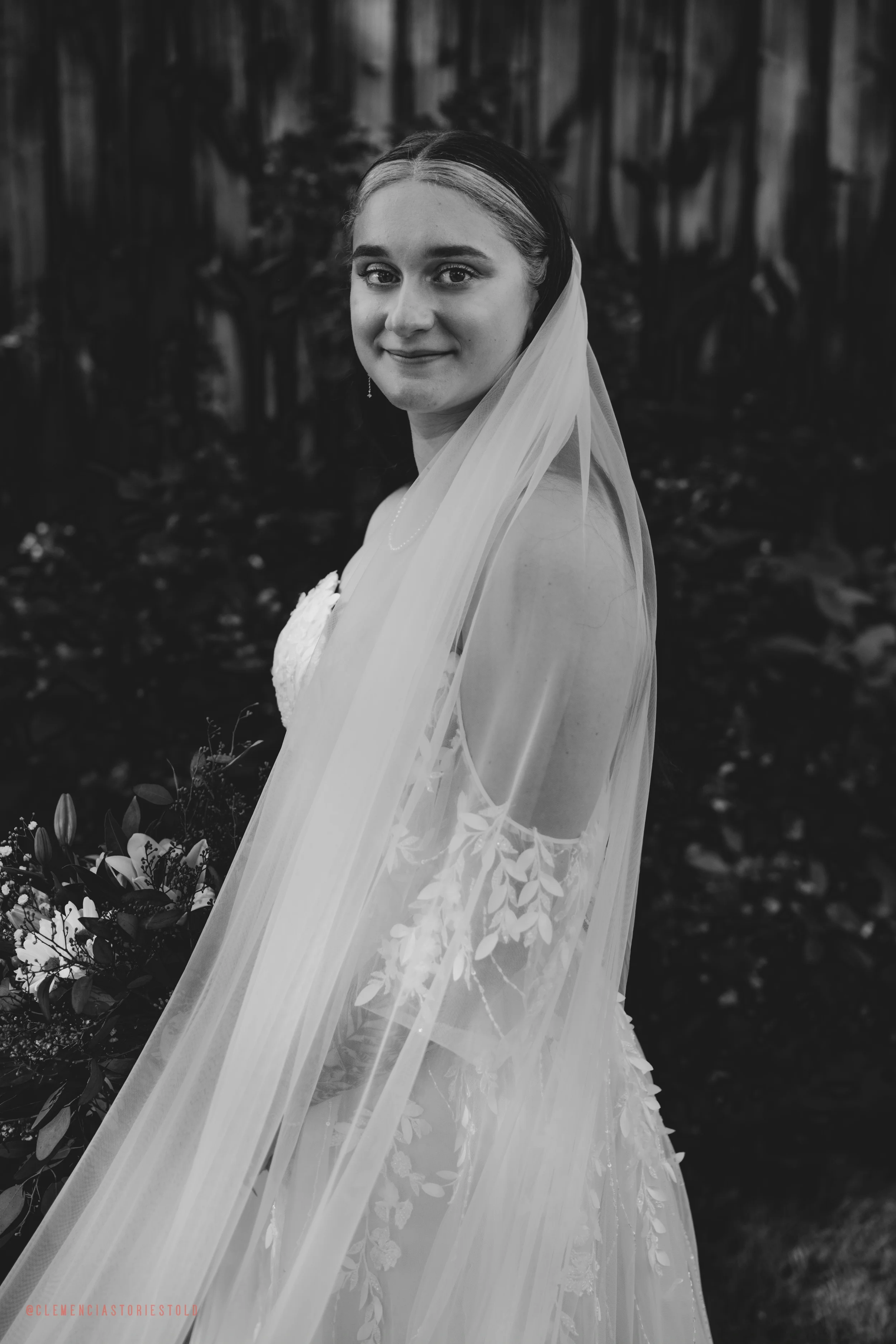 A bride in a wedding dress with a veil, standing outdoors with a wooden fence and trees in the background, holding a bouquet of flowers.
