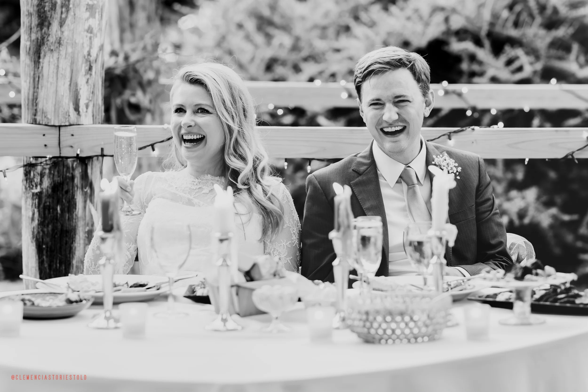 A joyous bride and groom laughing and celebrating at their wedding reception, seated at a table decorated with candles, glasses, and food, outdoors with string lights in the background.