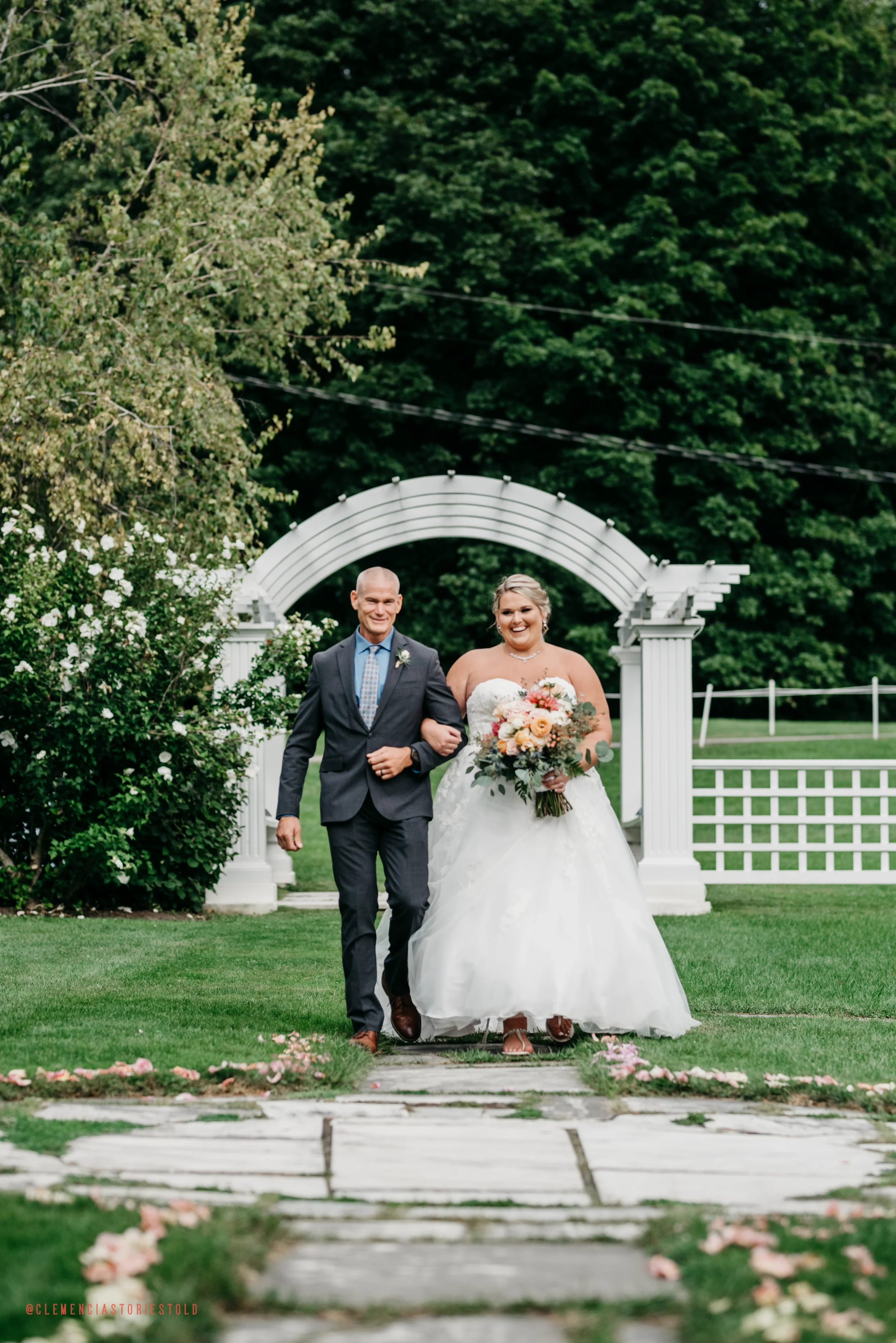 A bride in a white wedding gown holding a bouquet of flowers, walking arm-in-arm with an older man in a suit, outdoors on a grassy wedding path with a white arch and lush green trees in the background.