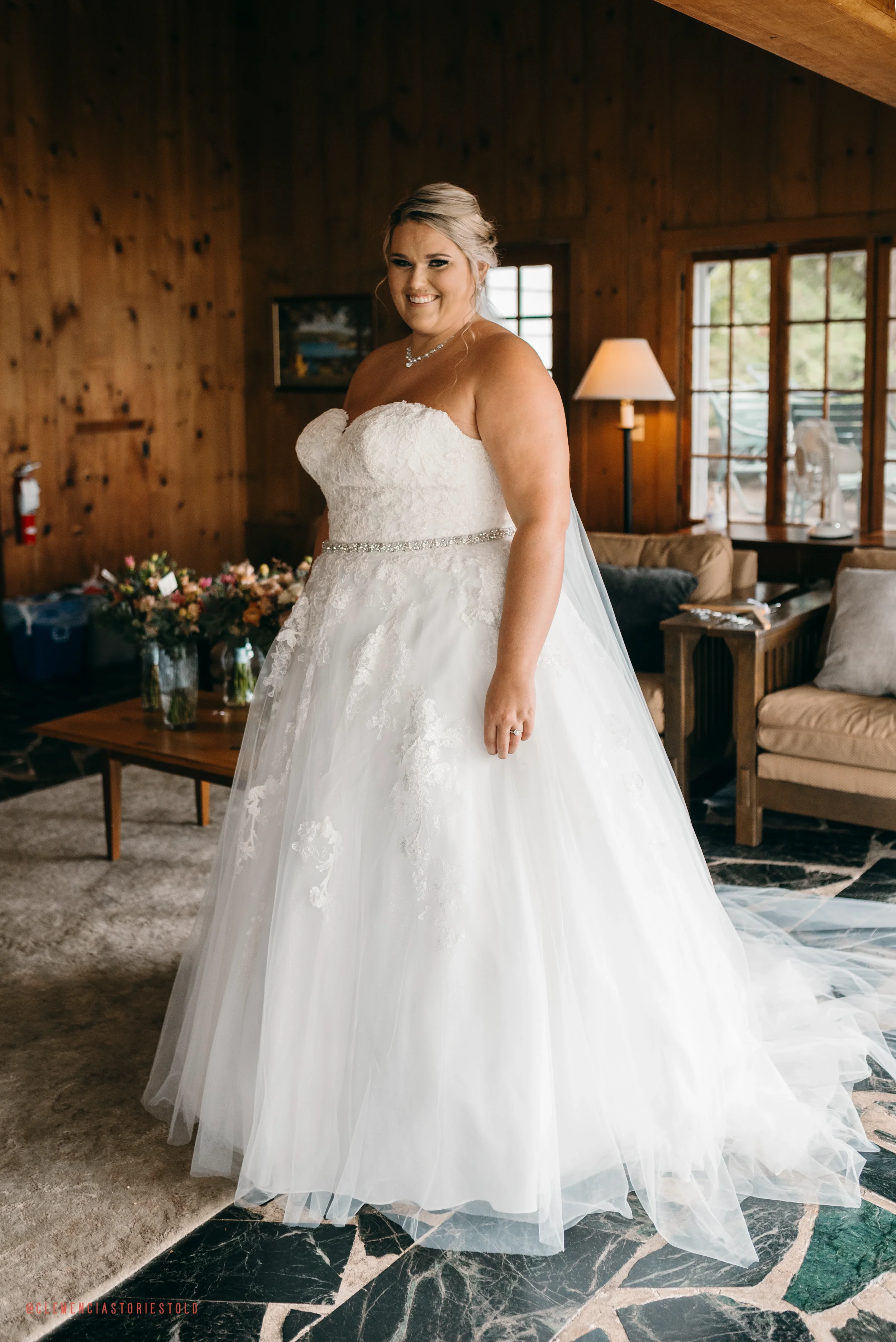A bride wearing a white wedding gown with a strapless lace bodice and tulle skirt, standing inside a wood-paneled room with large windows, a lamp, and a floral bouquet on a table in the background.