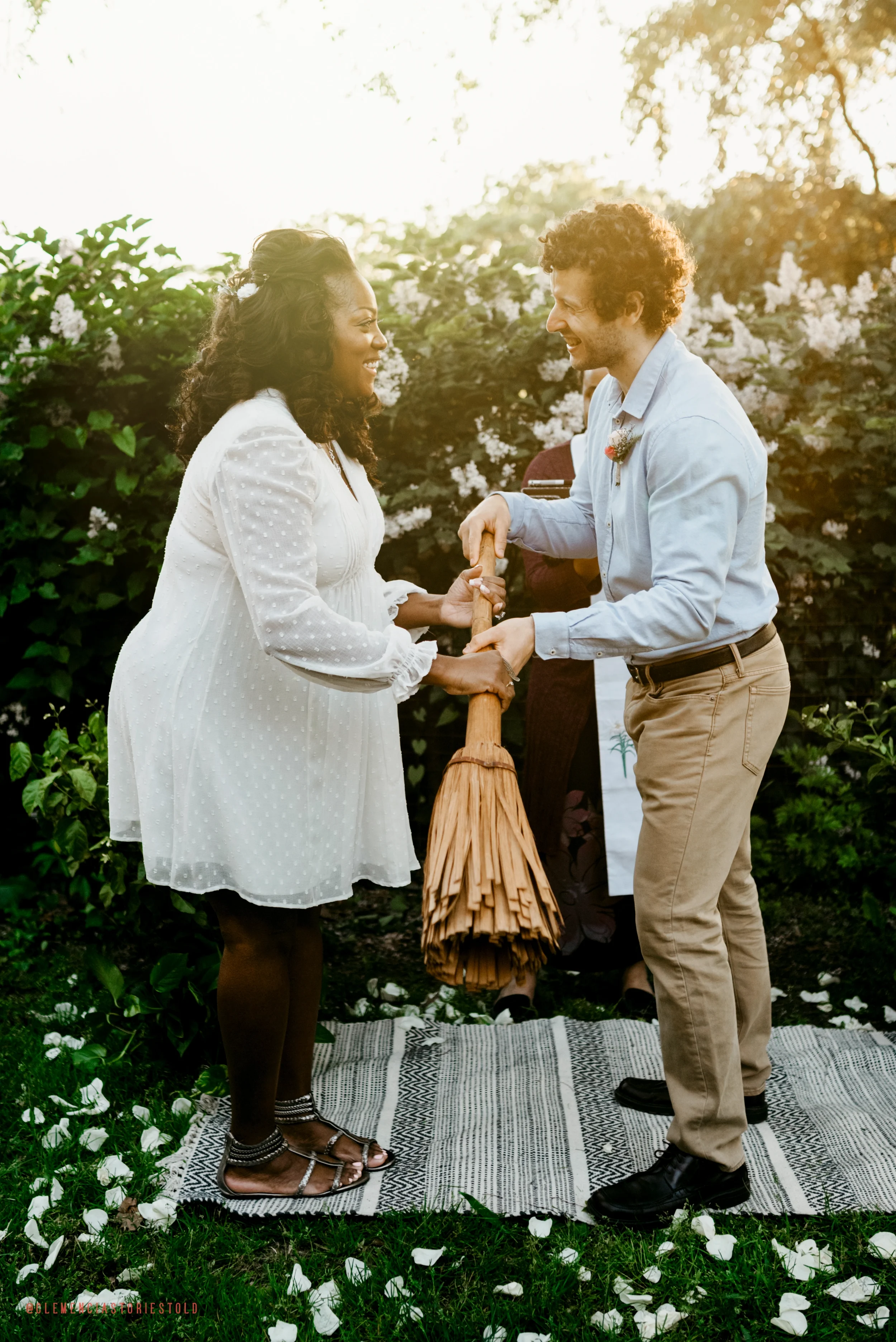 A couple holding hands during a wedding ceremony outdoors at sunset, with a woman officiant in the background and flower petals scattered on the ground.