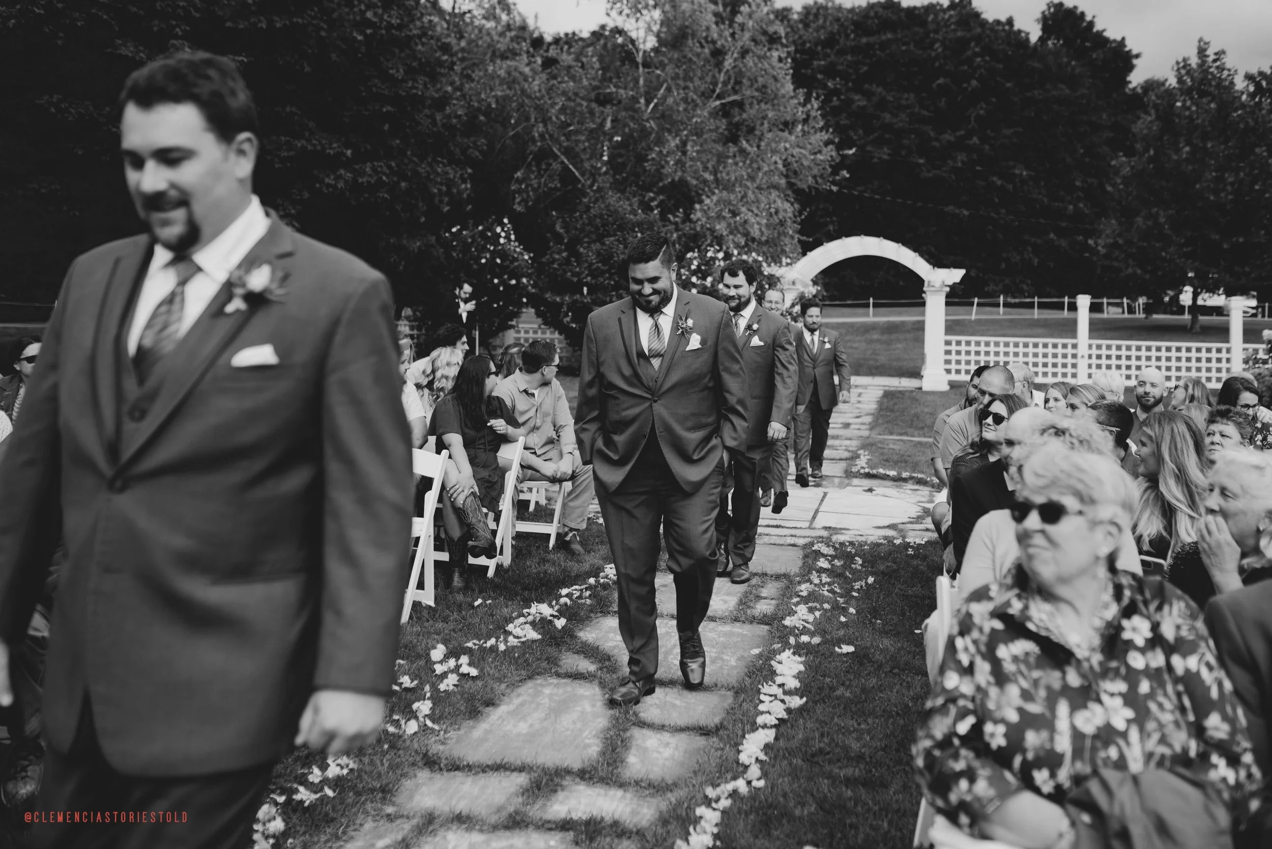 A group of men in suits walking down an outdoor aisle lined with flower petals, surrounded by seated guests at a wedding ceremony in a garden setting.