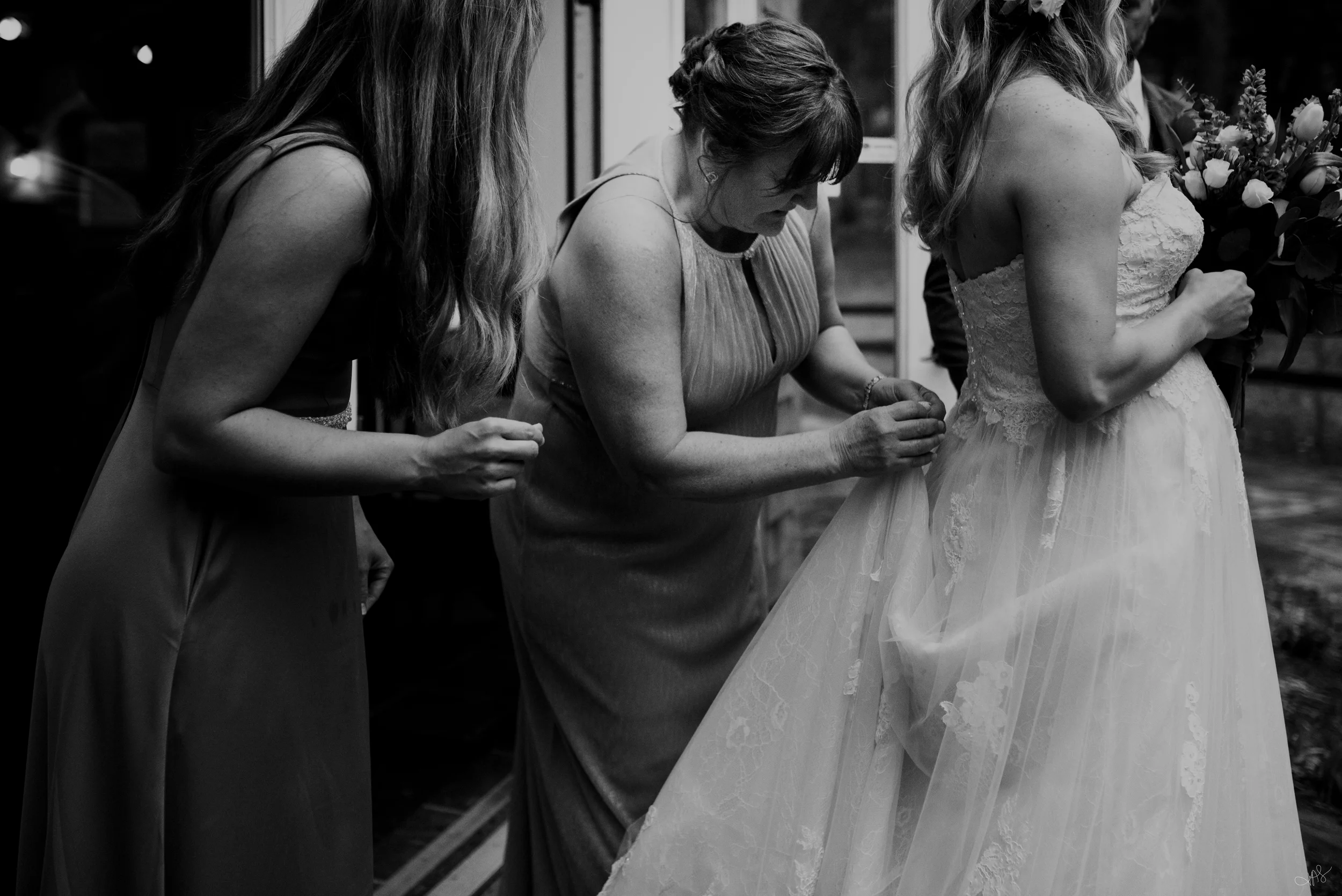 Women dressed in formal attire, including a bride in a wedding gown, gathered around and holding hands in a prayer circle during a wedding ceremony.