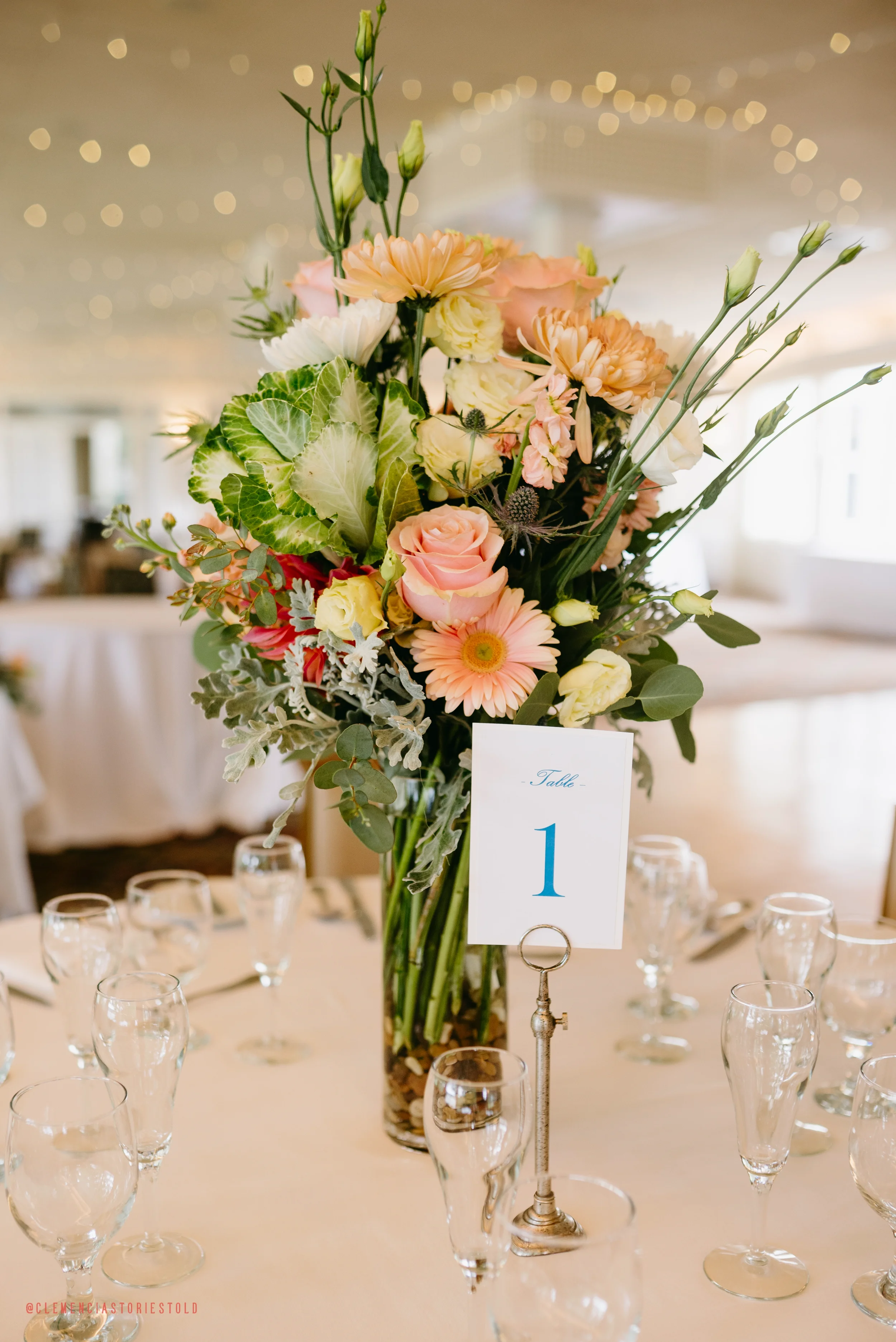A tall flower arrangement on a dining table with a table number sign. The arrangement includes pink roses, peach daisies, white lisianthus, green foliage, and other flowers. The table is set with multiple wine glasses and silverware.