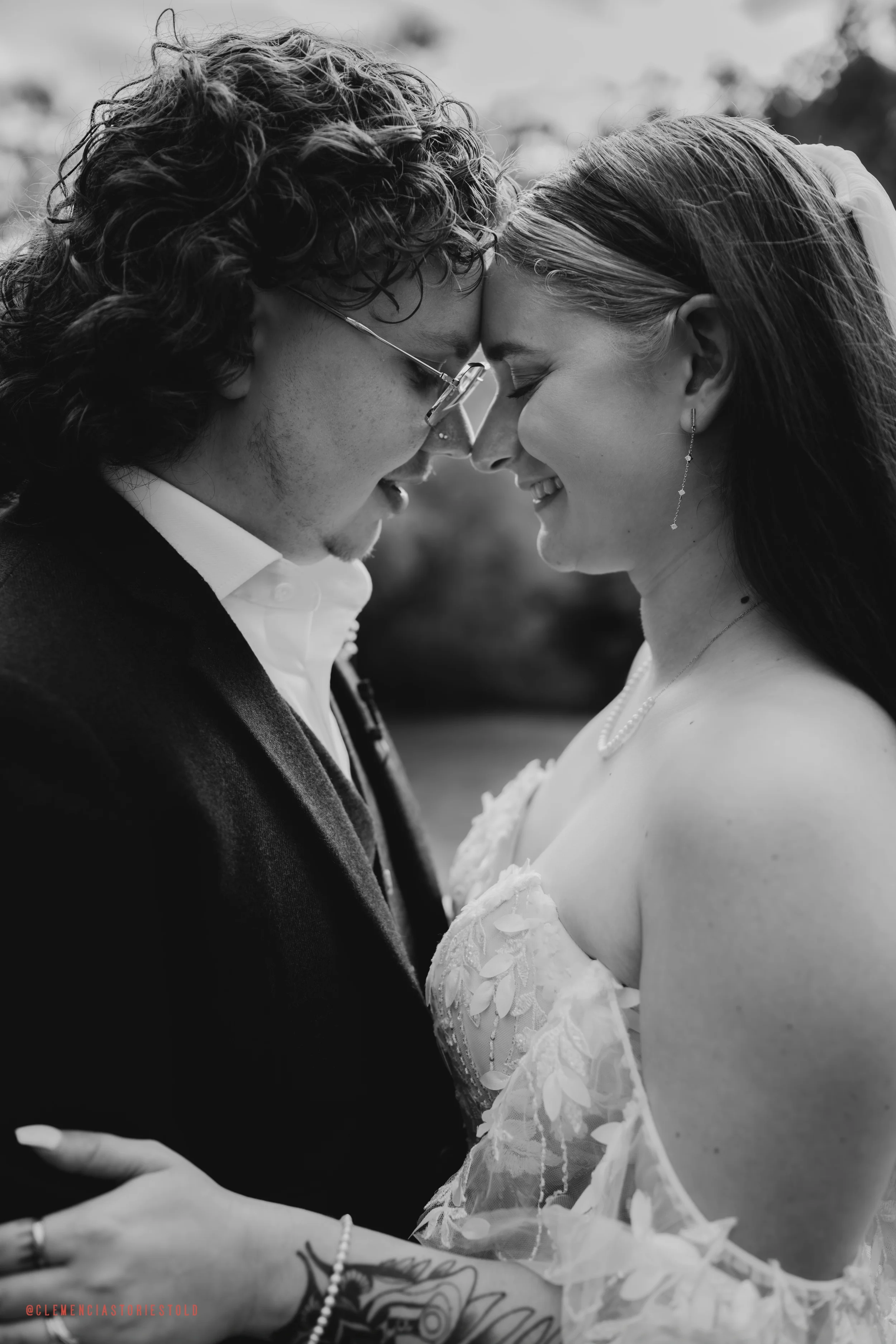 Black and white photo of a couple, a man and a woman, with their foreheads touching and eyes closed, smiling. The woman is wearing a lace wedding dress and the man is in a suit. They are outdoors with blurred trees in the background.