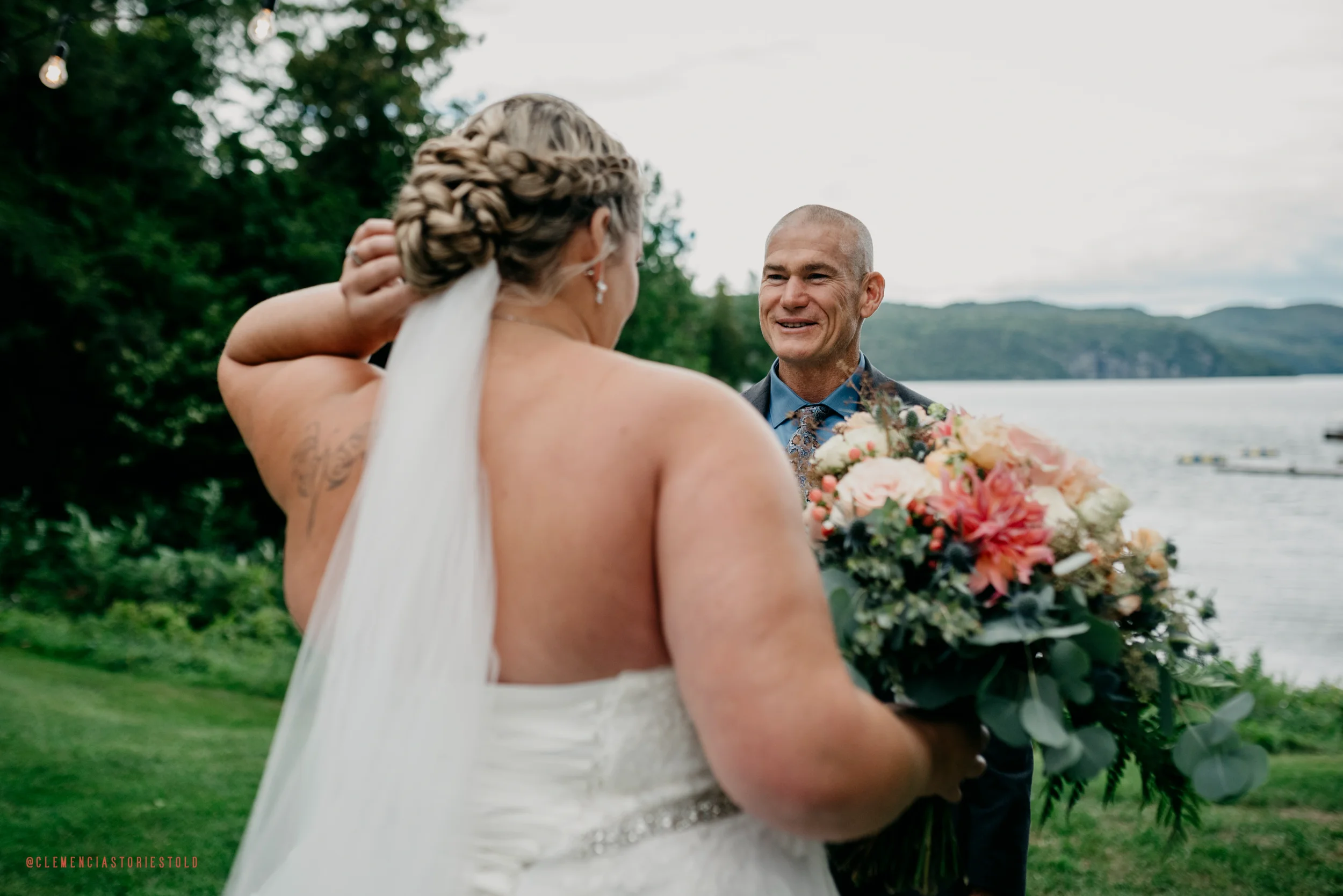 A bride with braided hairstyle holding a bouquet of flowers, smiling at her groom by a lake with wooded hills in the background.