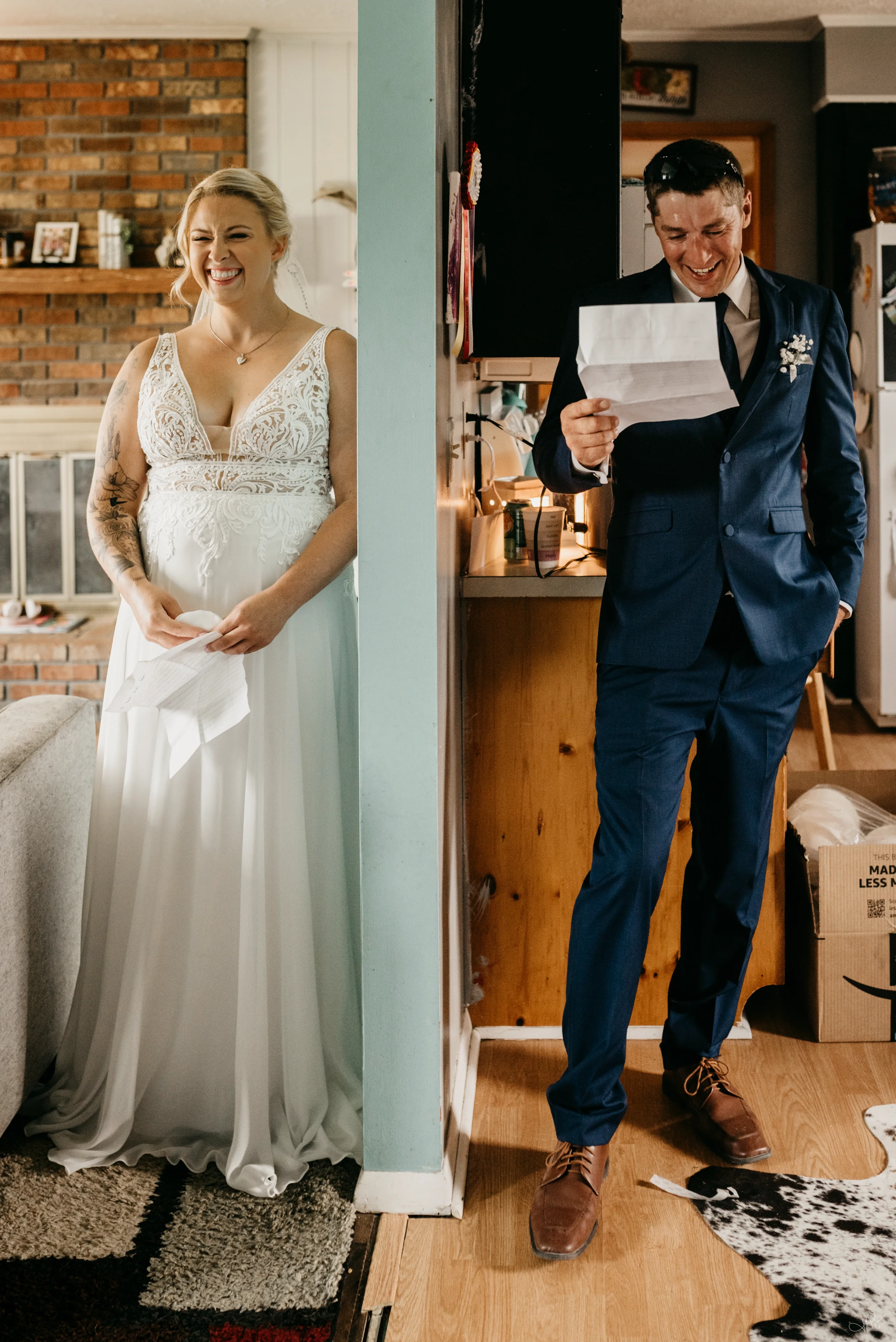 A bride and groom at their wedding, standing on opposite sides of a wall. The bride is smiling in her white dress with tattoos on her arms, and the groom is smiling while reading a paper, dressed in a blue suit with a boutonniere.