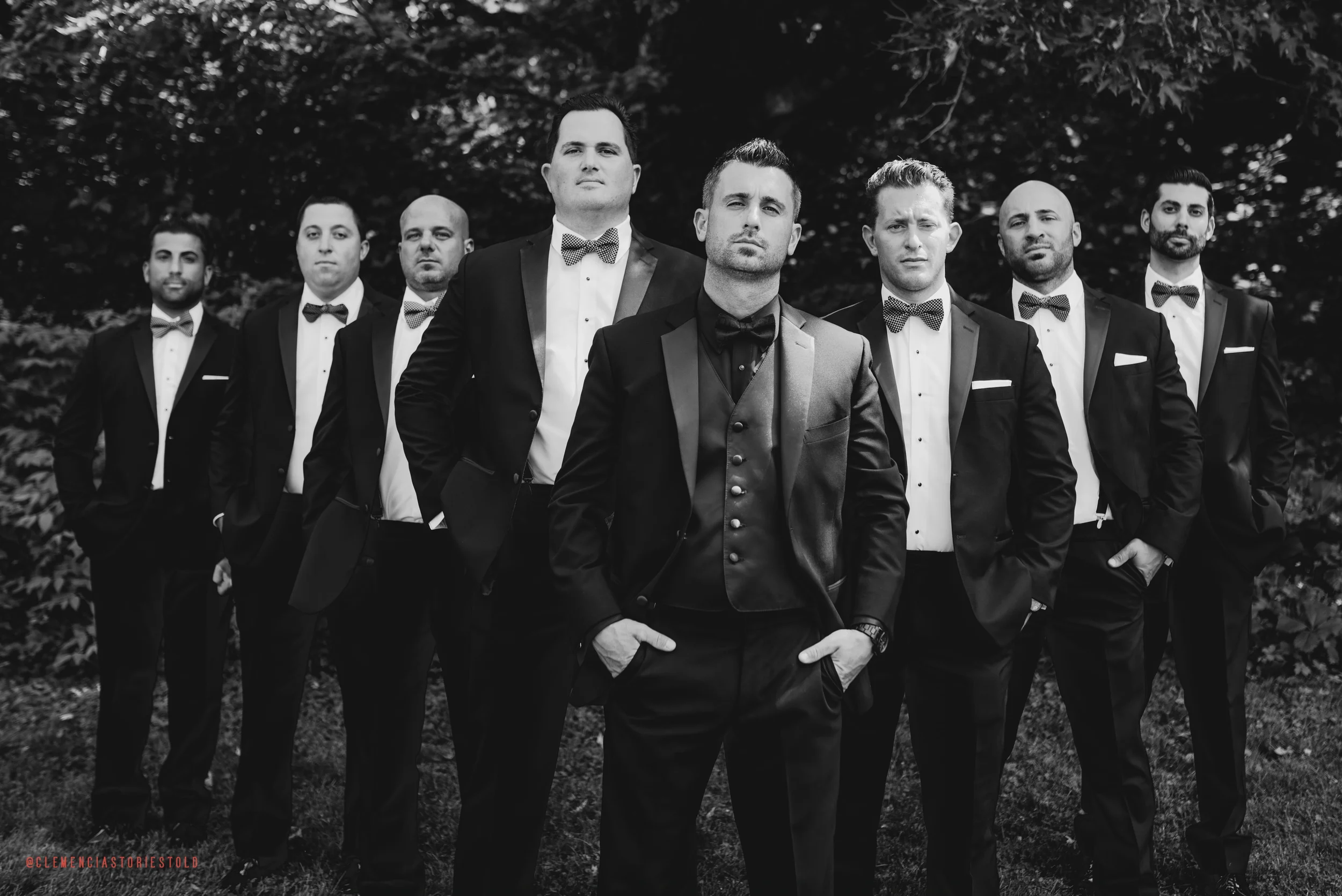A black and white photograph of nine men dressed in formal tuxedos with bow ties, standing outdoors in front of trees, with serious expressions and confident postures.