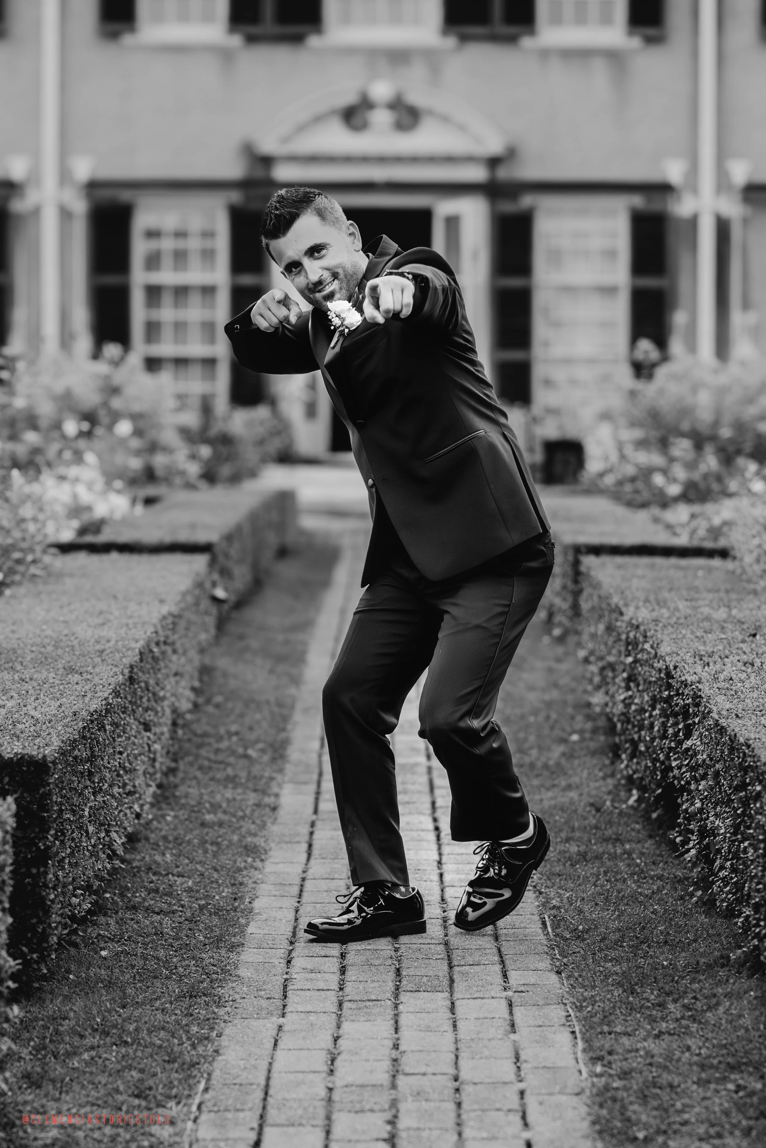 Black and white photo of a man in a suit standing on a brick pathway, pointing towards the camera, with a building and garden in the background.