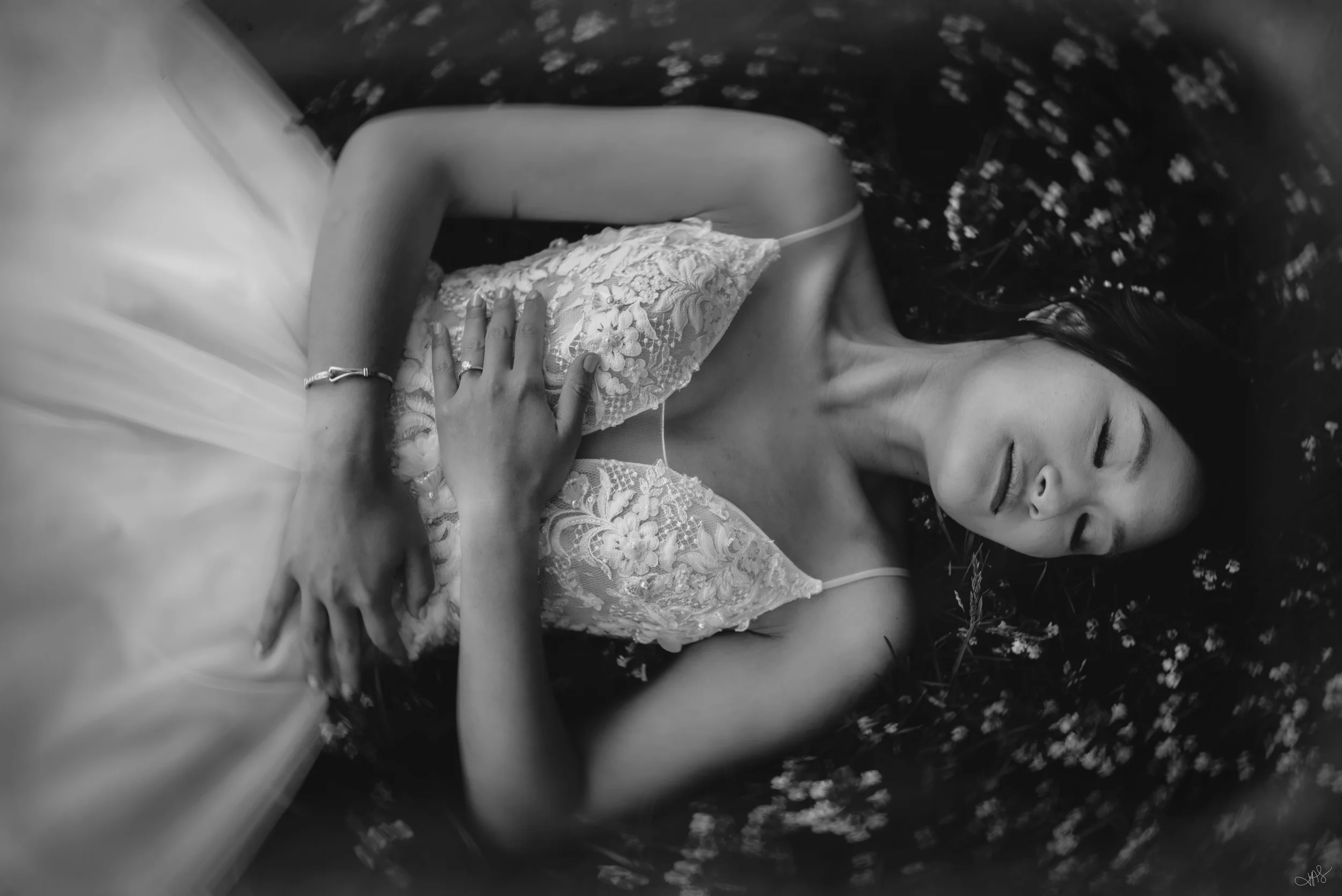 A woman relaxing outdoors on her back, wearing a lace top with floral embroidery, holding her stomach softly, with her eyes closed and a serene expression. She is lying on grass with small flowers around.