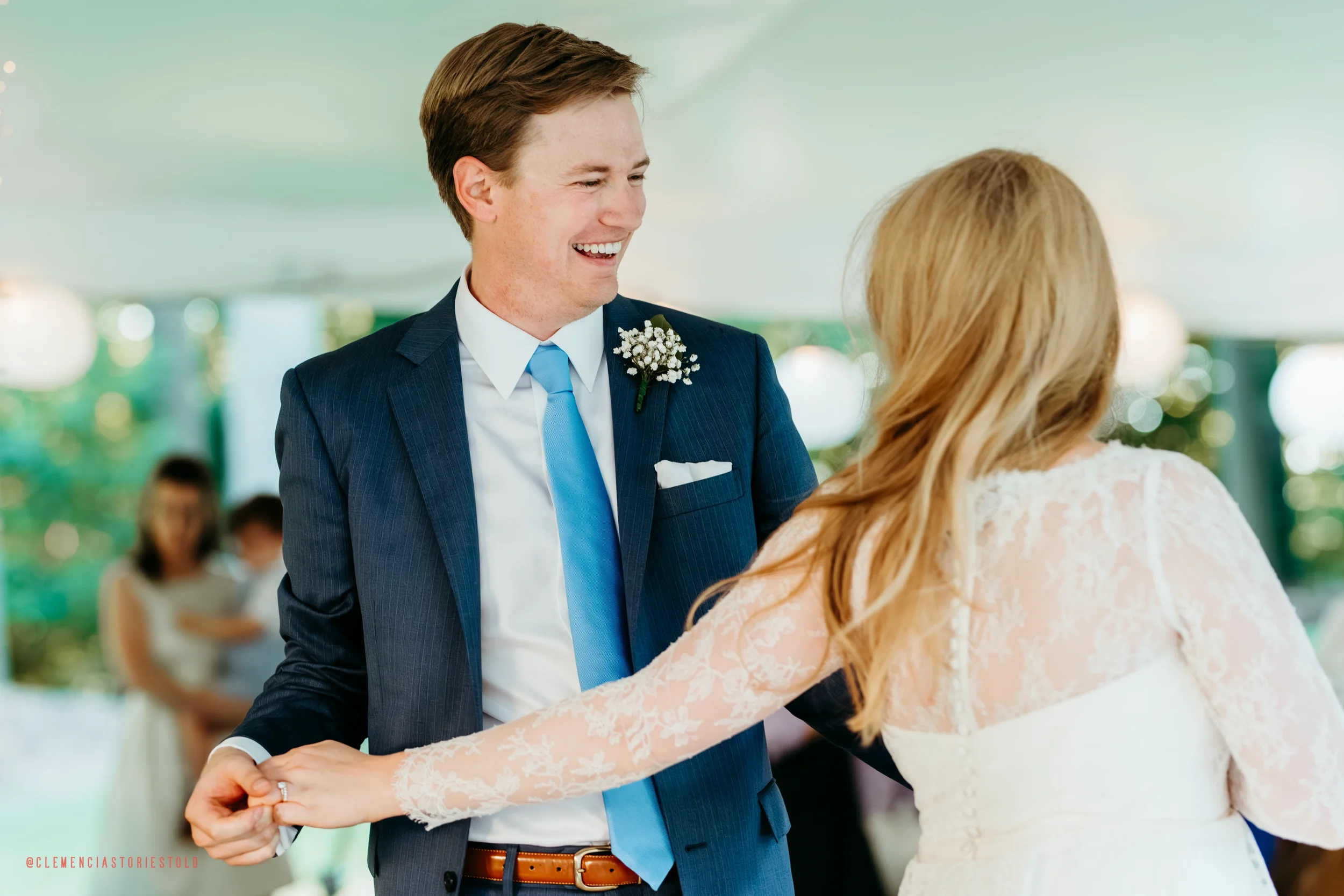 A bride and groom dancing at their wedding reception, holding hands and smiling, with guests in the background.