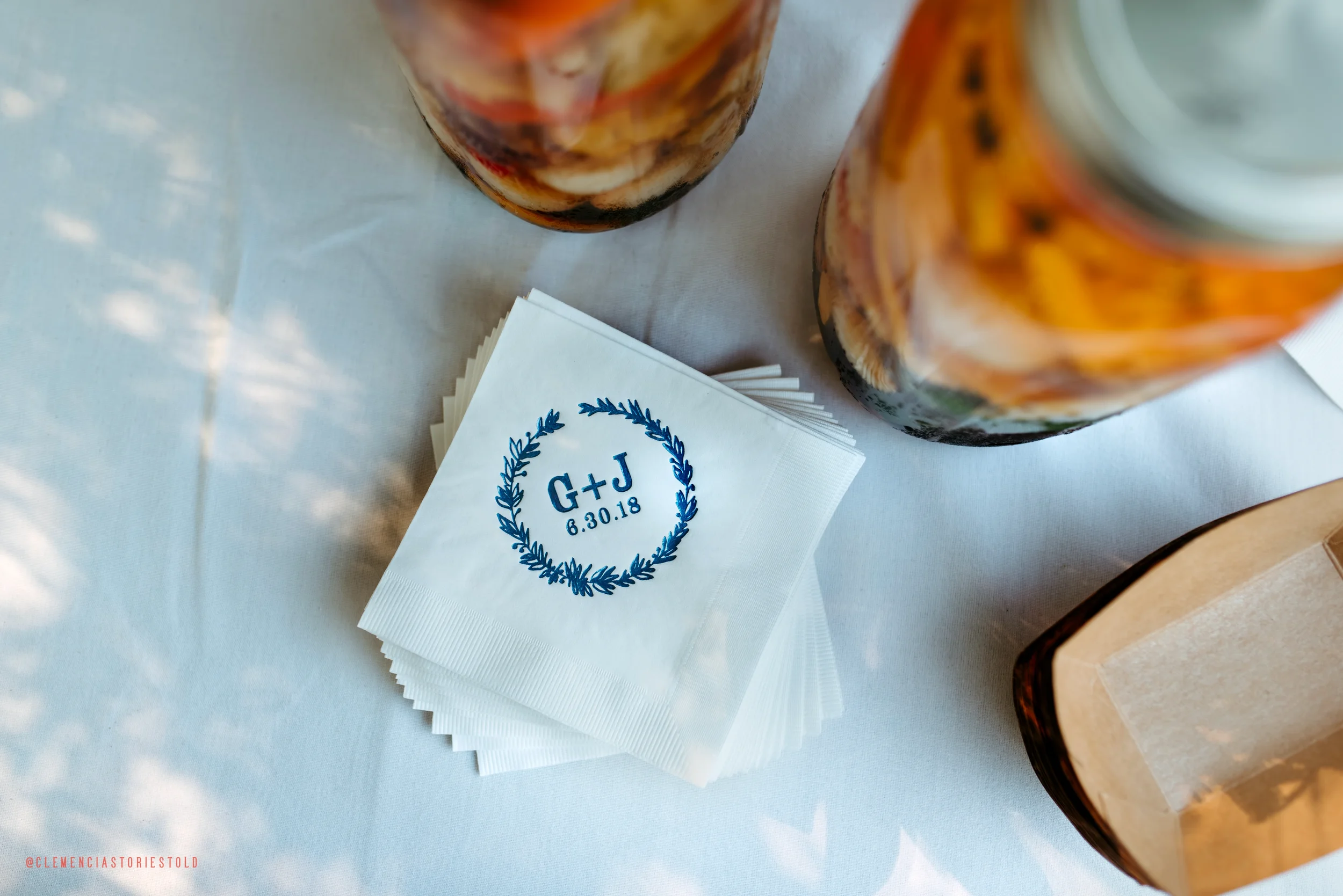 Wedding napkin with initials G+J and the date 8.30.18, placed on a white tablecloth, surrounded by glass jars and a small box.