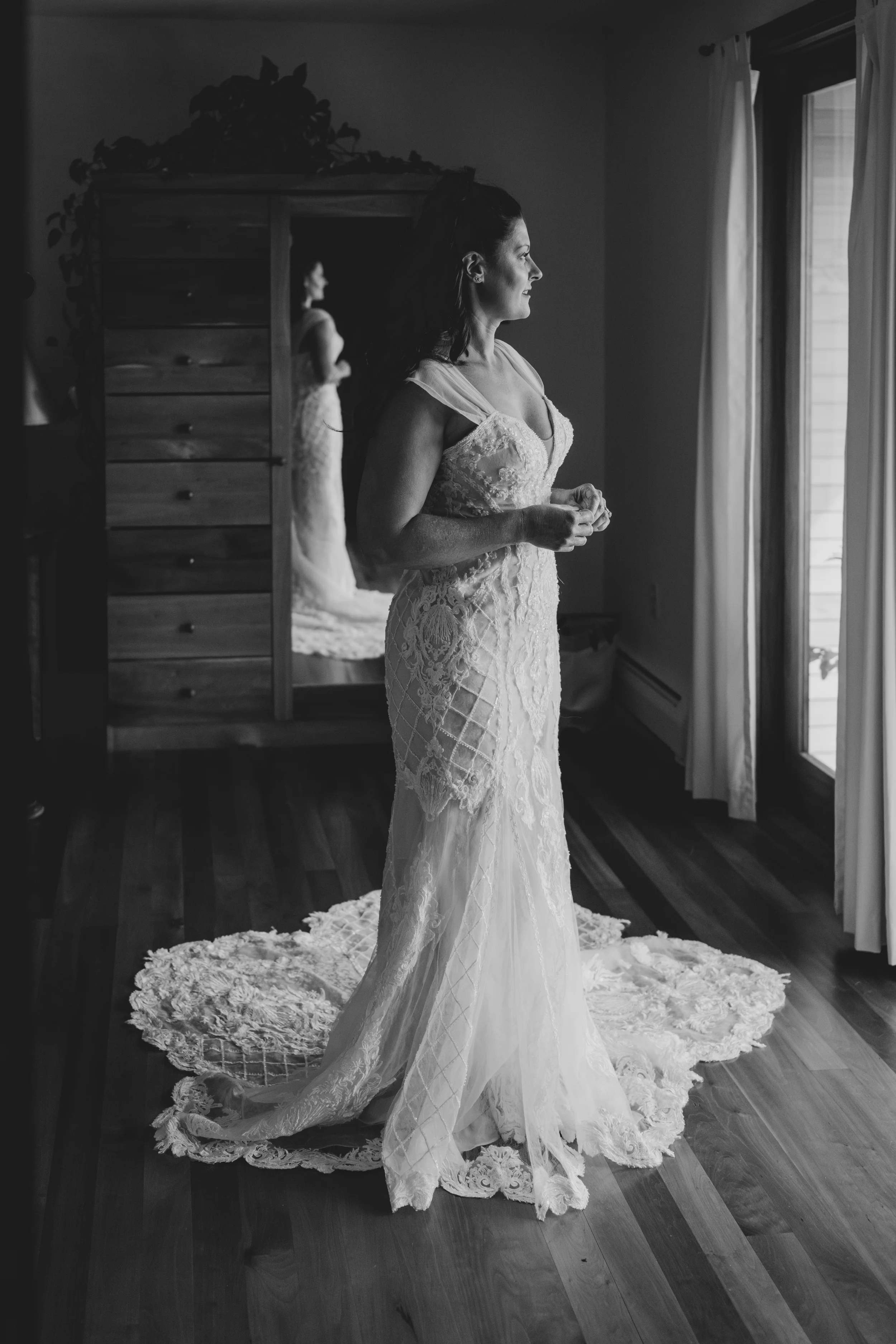 A woman in an elegant wedding dress stands in a room, looking out a window with curtains, with a large floral rug on the wooden floor and a dresser with a mirror behind her.