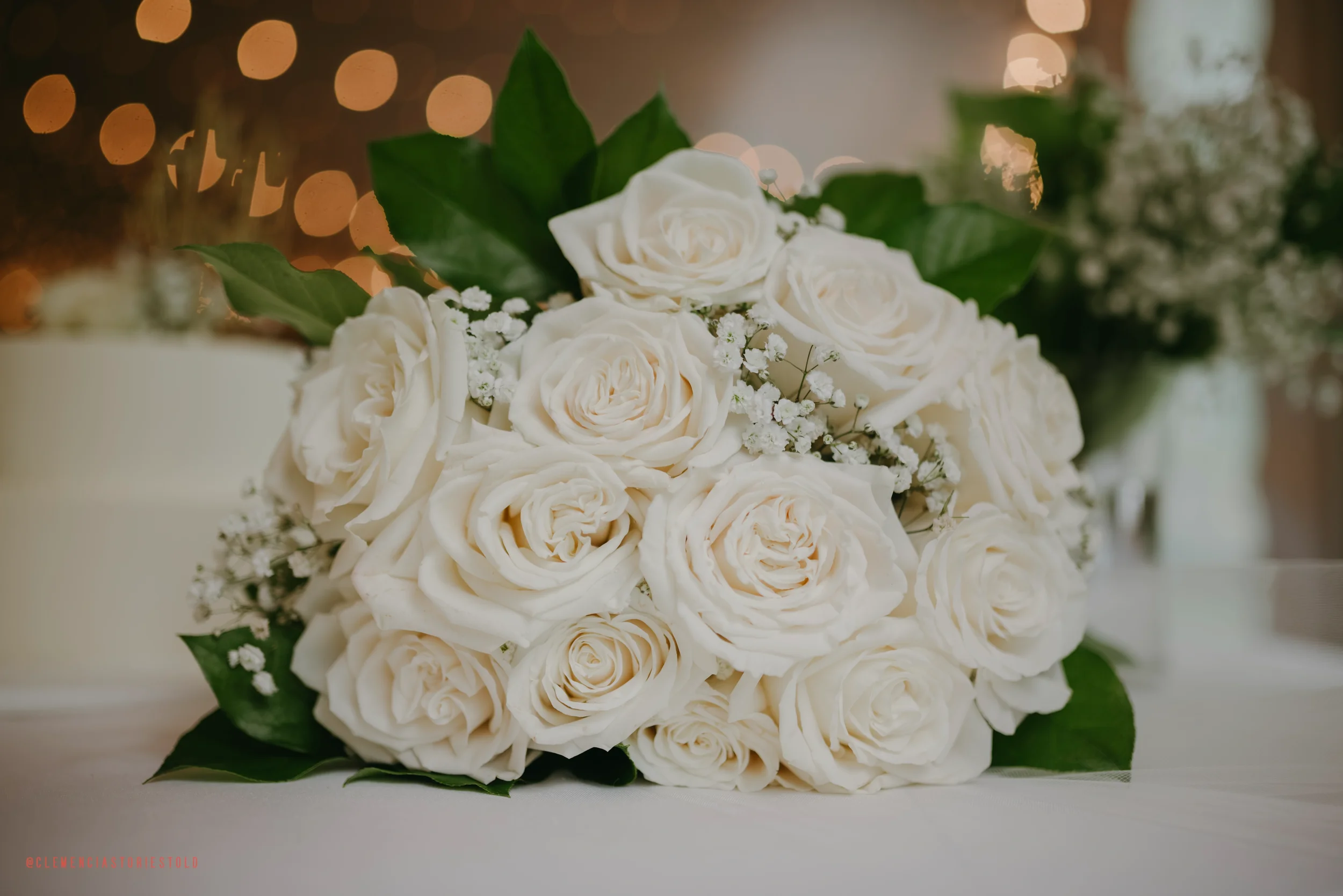 A bouquet of white roses with green leaves and small white filler flowers, placed on a white surface with a blurred background of warm lights.