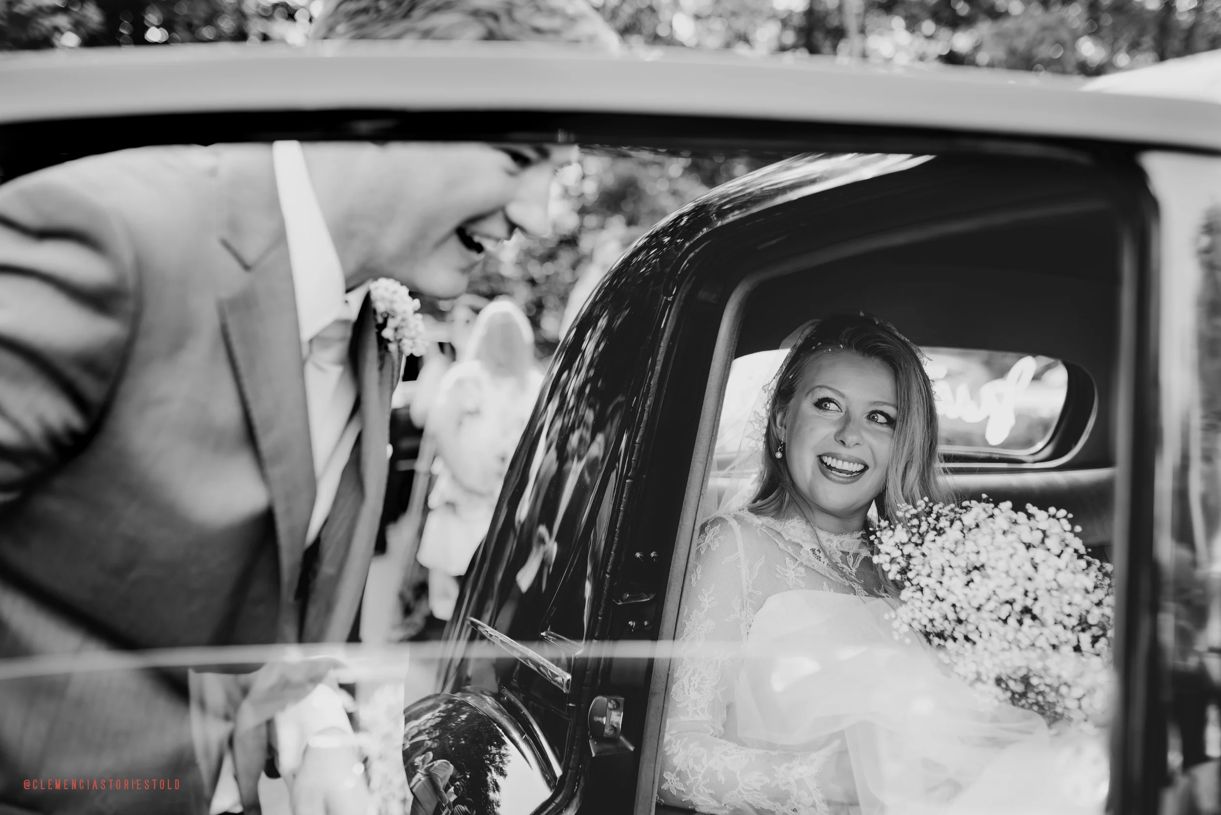 Black and white photo of a bride sitting in a vintage car, smiling at a man leaning in from outside, with flowers in her hand.