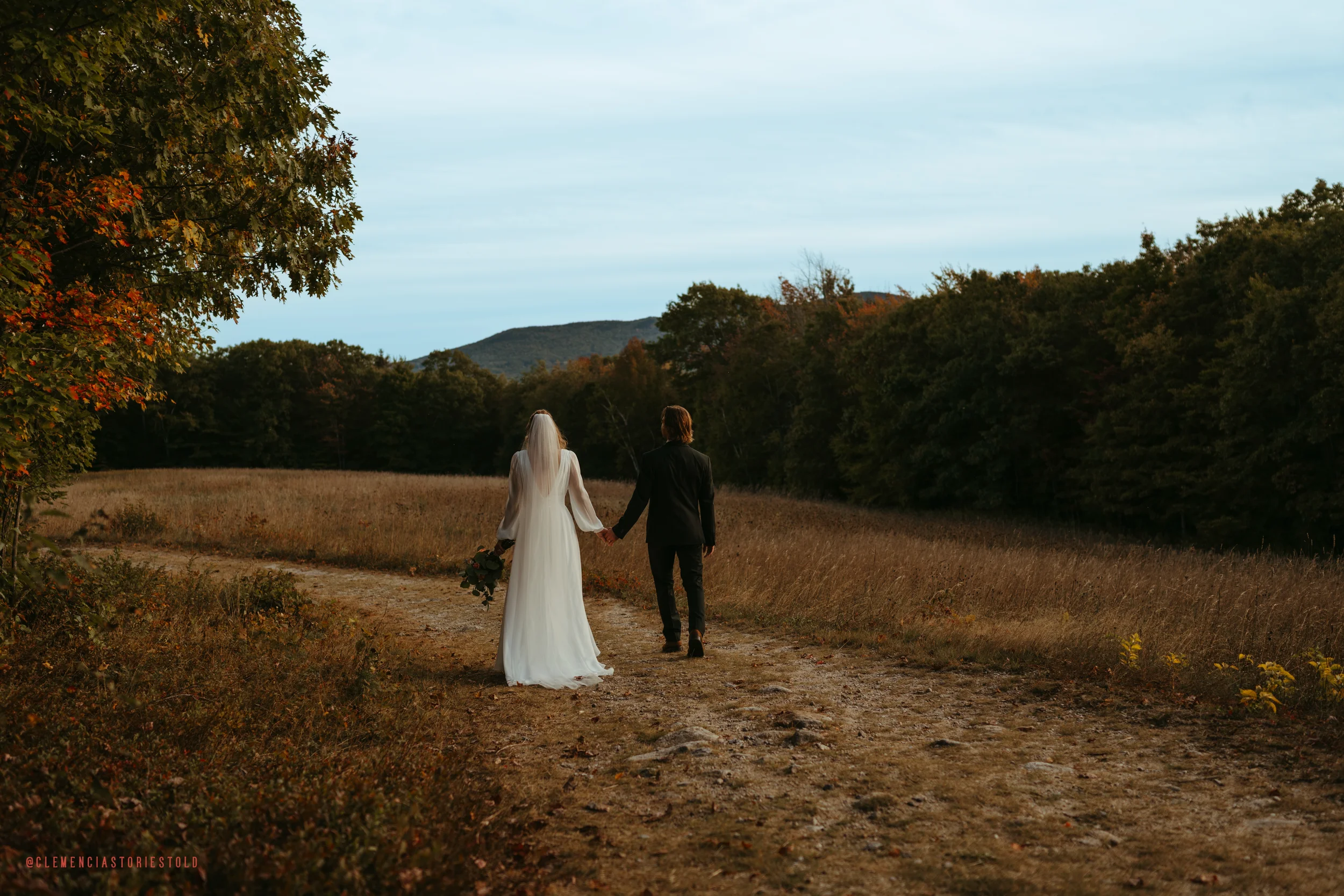 A bride and groom walk hand in hand along a dirt path through a natural landscape with trees and mountains in the background during sunset.