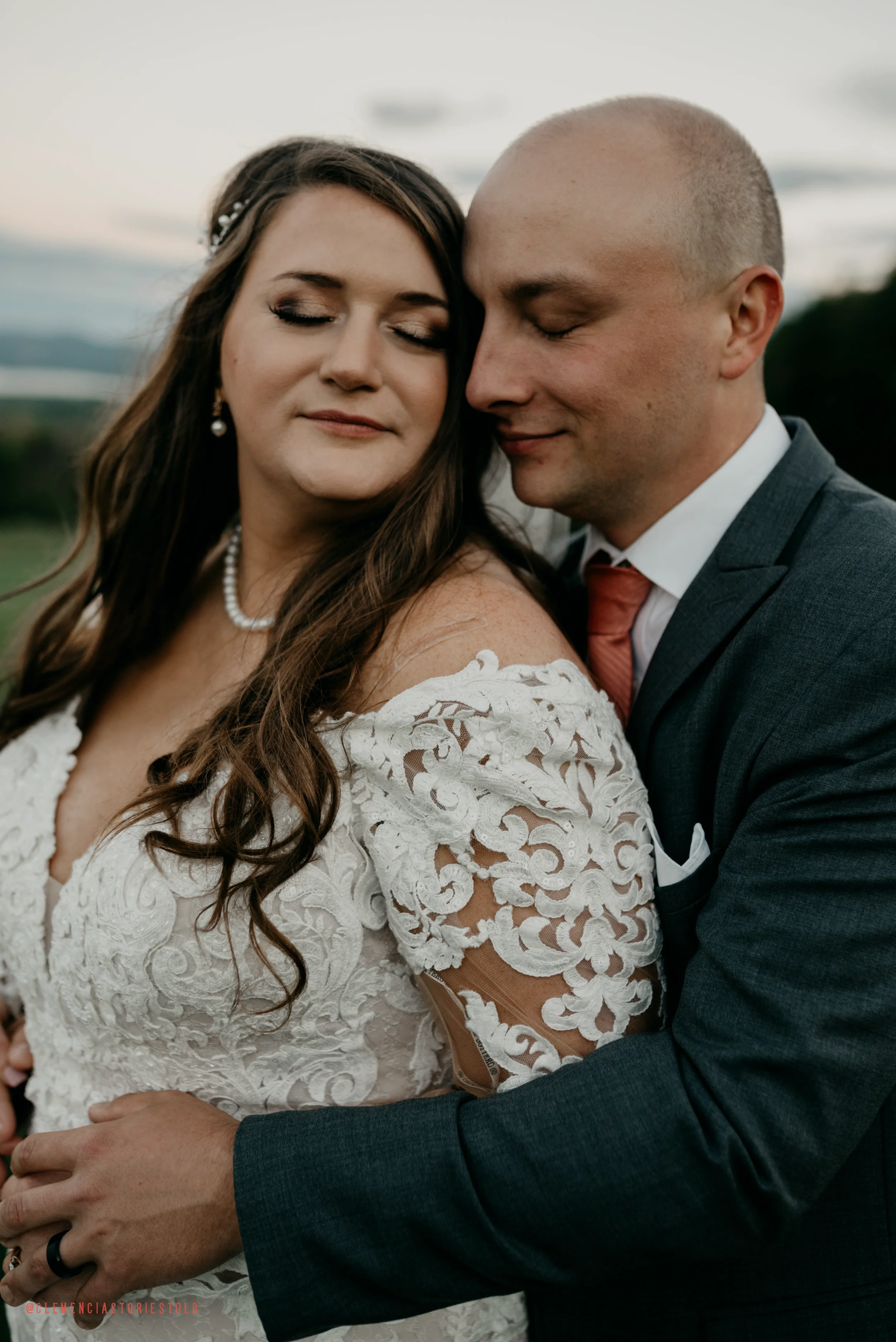 A bride and groom with close eyes, embracing outdoors during sunset, with a scenic landscape in the background.