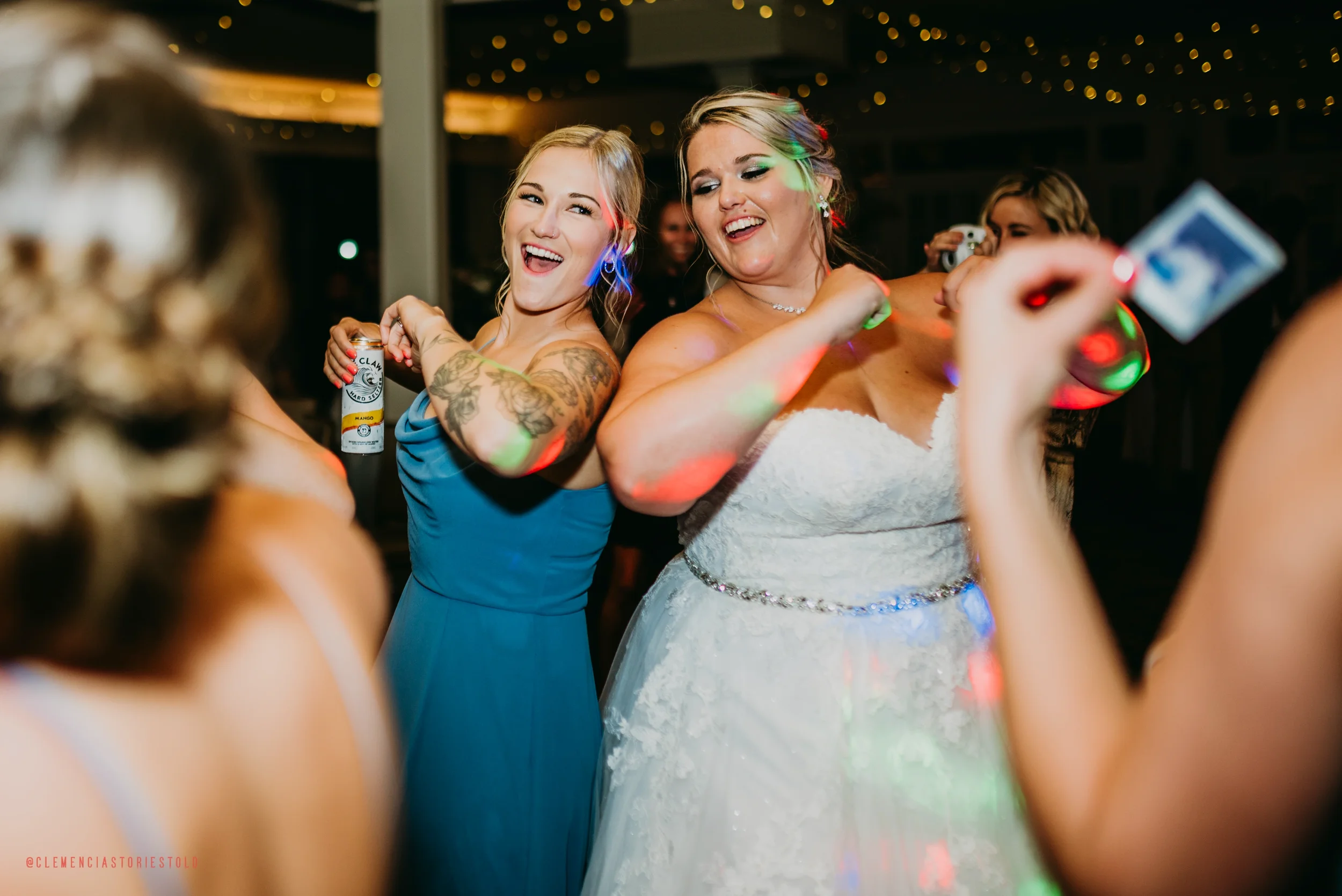 Two women, one in a wedding dress and the other in a blue gown, are dancing and smiling at a celebration with colorful lights.
