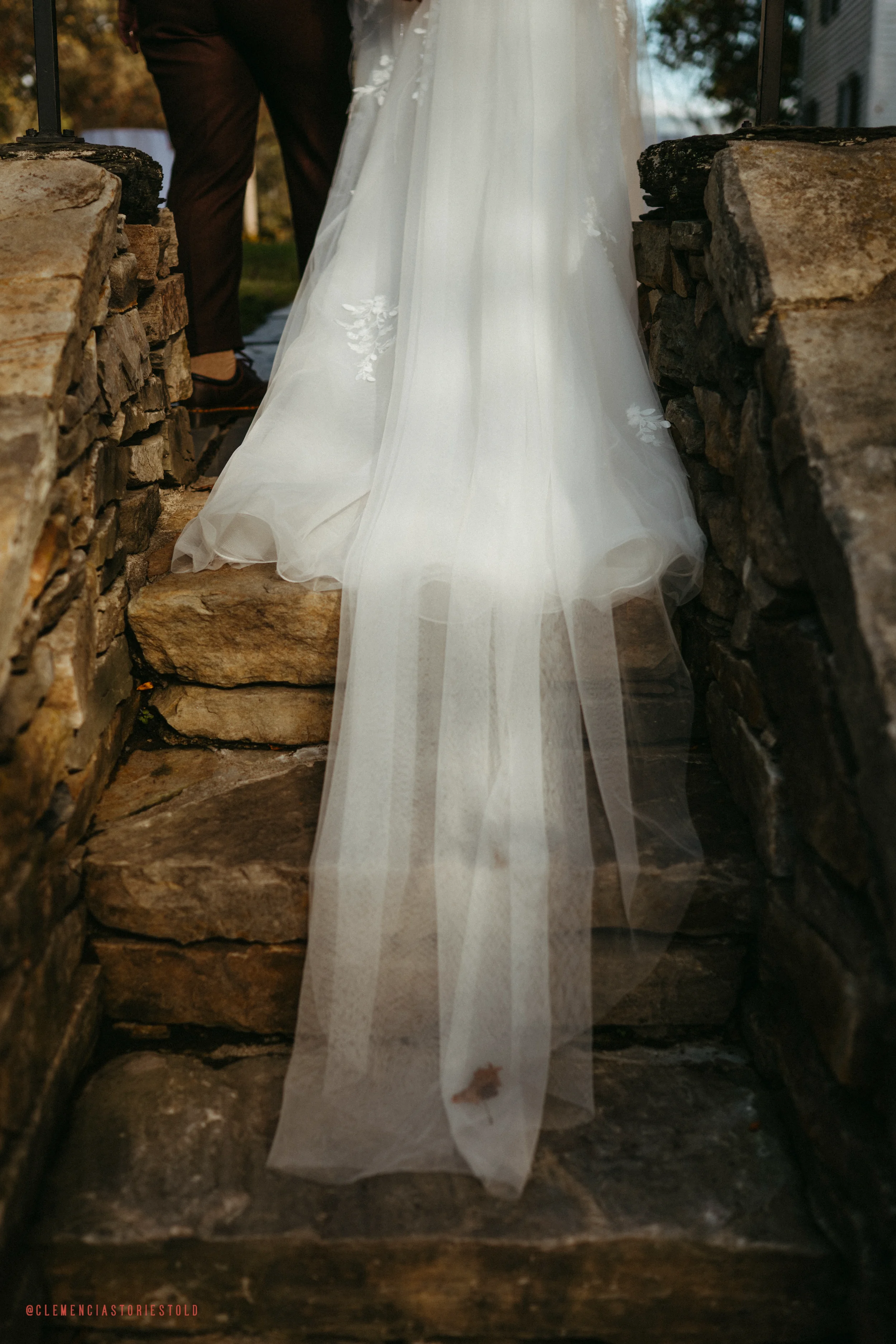Close-up of a bride's white wedding gown with a tulle train, standing on rustic stone steps between stone walls.