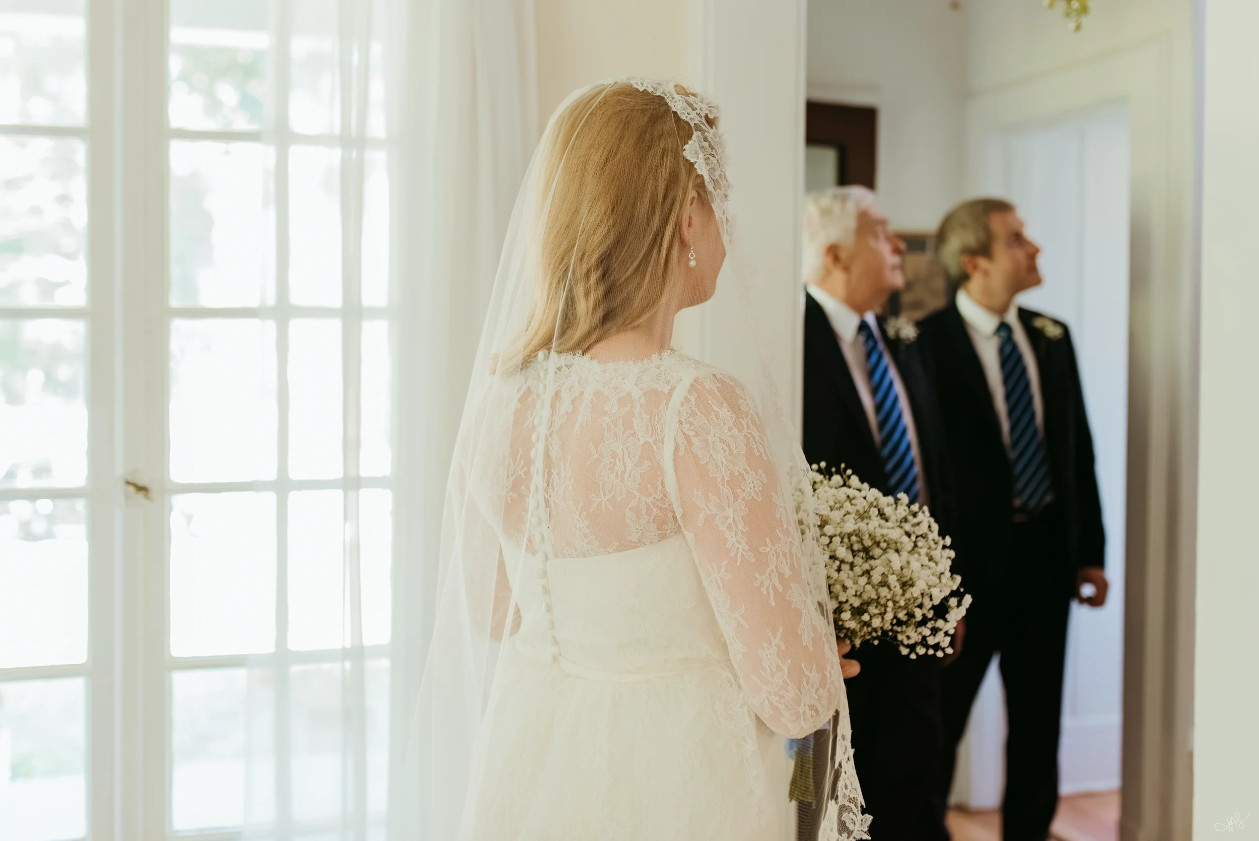 A bride in a lace wedding dress with a veil and earrings stands indoors, holding a bouquet of white flowers. Two men in suits stand in the background near a large window with white curtains.