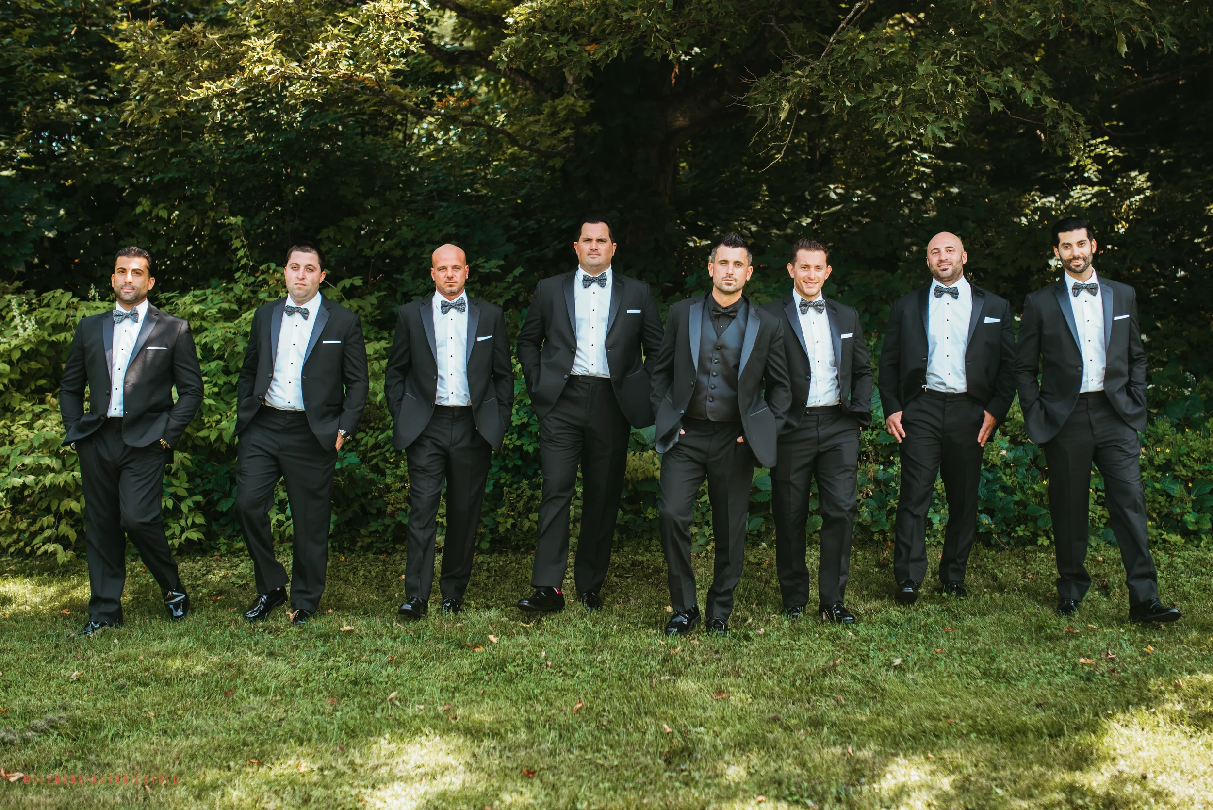 A group of nine men dressed in black tuxedos with white shirts and bow ties standing outdoors on a grassy area with green trees and bushes in the background.