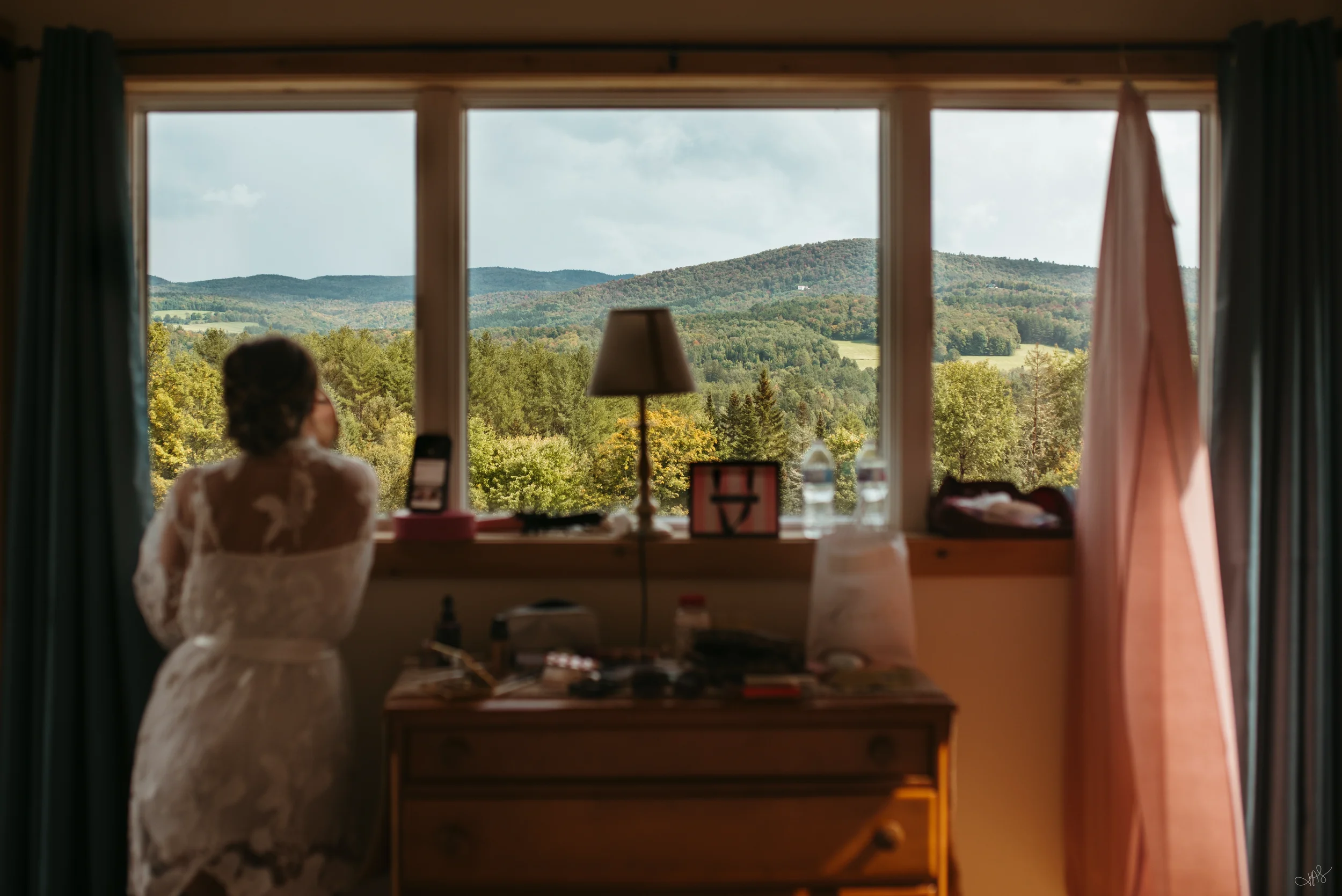 A person in a wedding dress sitting inside a room, looking out a large window at a scenic view of green hills and trees on a cloudy day.