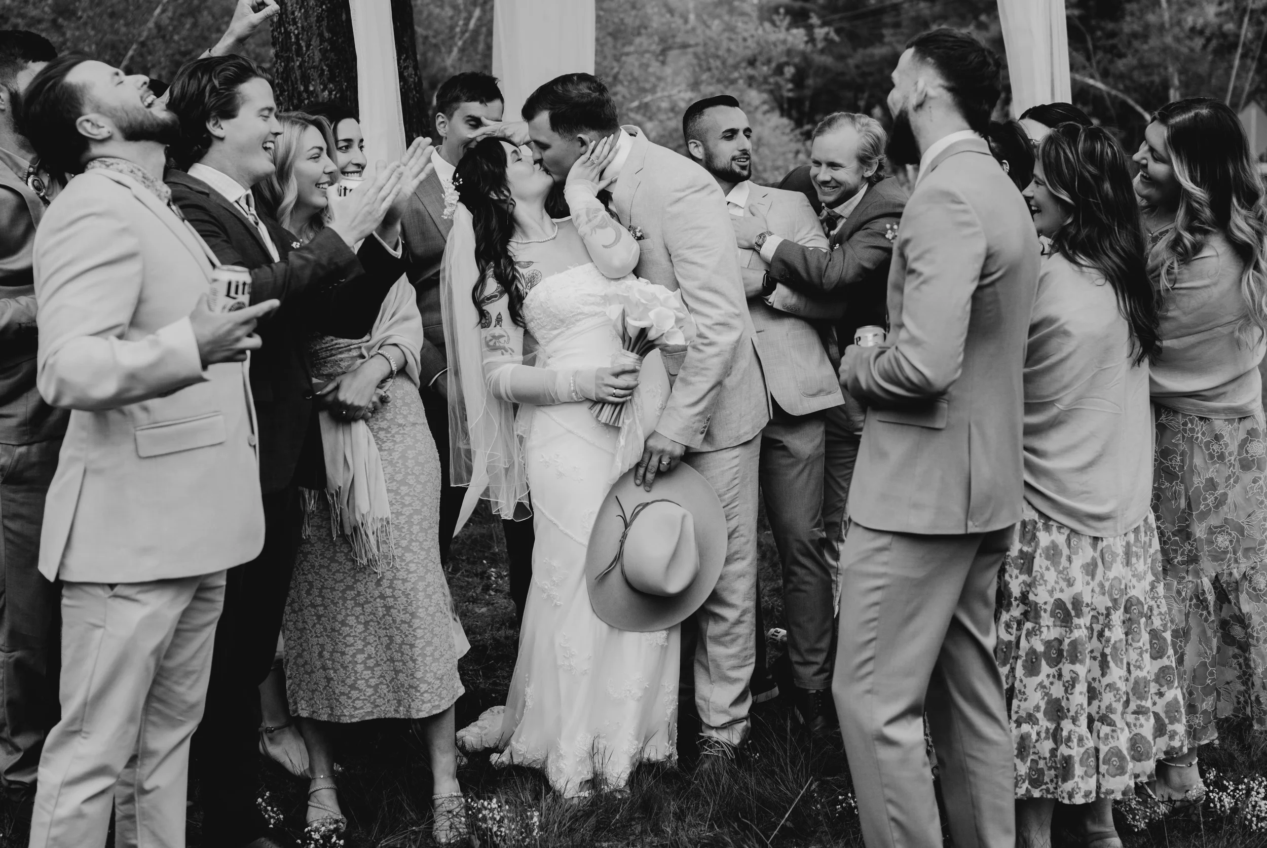 Black and white photo of a wedding scene with a bride and groom kissing among a group of people.