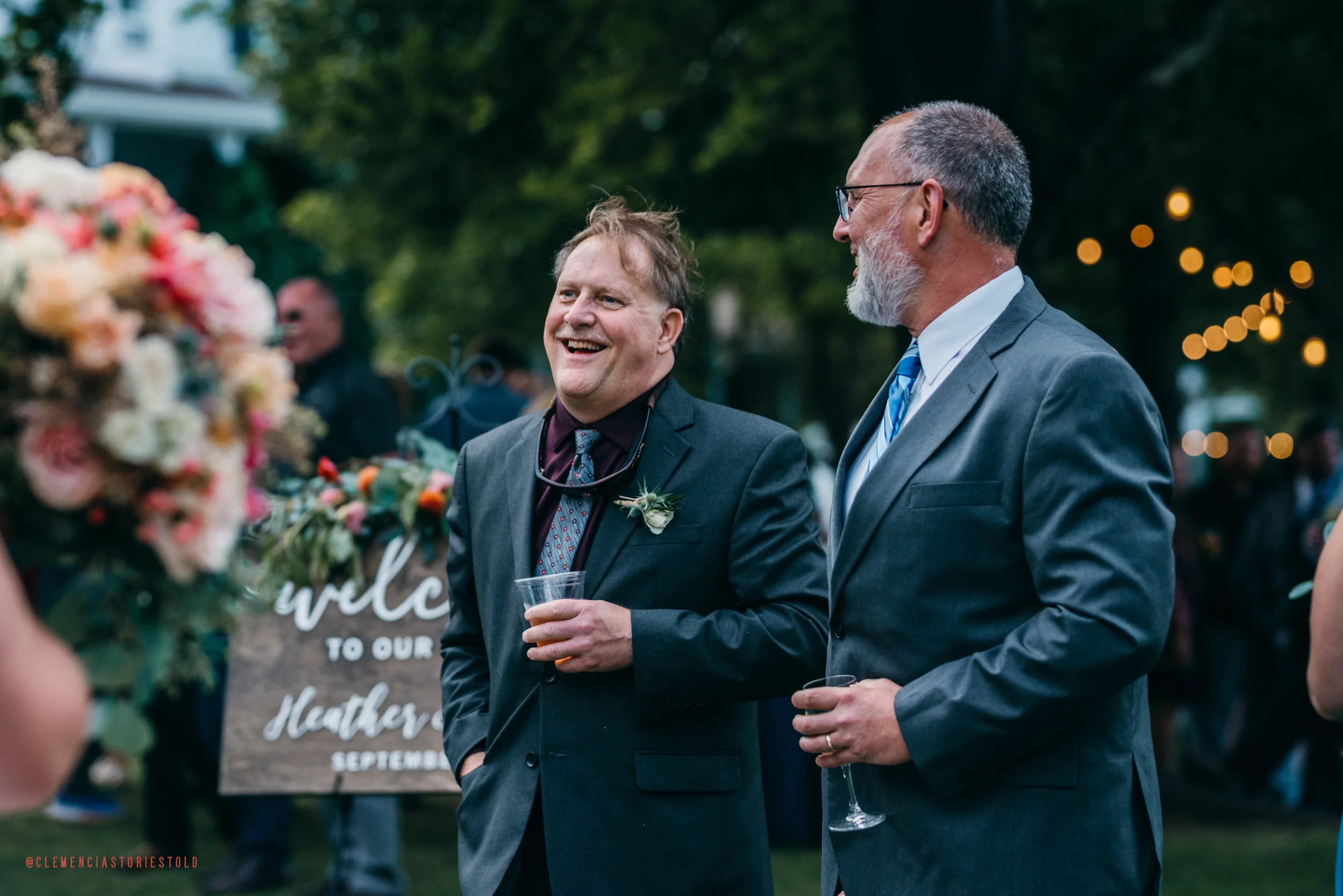 Two men in suits are smiling and talking at an outdoor wedding reception, holding drinks in their hands.